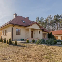 Widok z zewnątrz na parterowy dom z poddaszem użytkowym, z czerwoną dachówką, beżową elewacją, kamiennym wykończeniem wejścia i zadbanym ogrodem z iglakami oraz dodatkowym budynkiem gospodarczym...