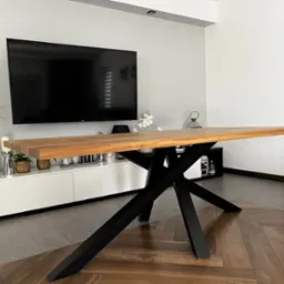 Stół z blatem z jasnego drewna i czarną, geometryczną podstawą w nowoczesnym salonie z telewizorem i dekoracjami.