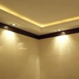 Wnętrze pomieszczenia z wykończonym narożnikiem sufitu, gdzie zamontowano oświetlenie LED w listwie przysufitowej oraz punktowe lampy halogenowe, tworzące ciepłą barwę światła na gładkich, beżowych...