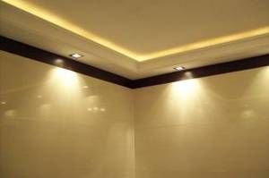 Wnętrze pomieszczenia z wykończonym narożnikiem sufitu, gdzie zamontowano oświetlenie LED w listwie przysufitowej oraz punktowe lampy halogenowe, tworzące ciepłą barwę światła na gładkich, beżowych...