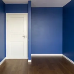 Pomalowane na granatowo wnętrze z drewnianą podłogą i białą stolarką drzwiową oraz listwami przypodłogowymi. Gładka, matowa powierzchnia ścian. Widoczne gniazdko elektryczne.