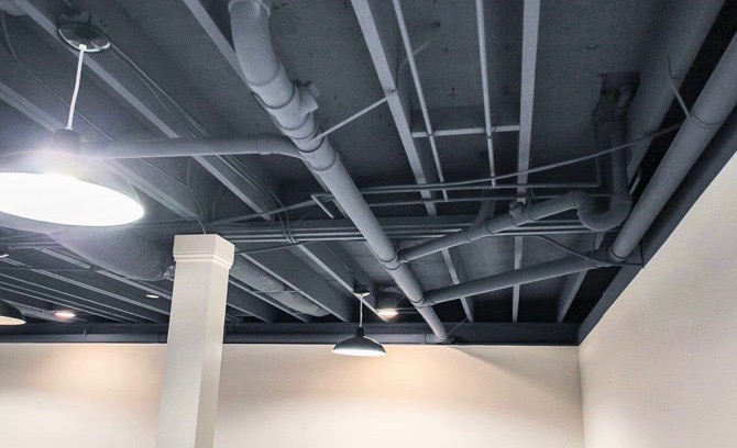 Pomieszczenie z odsłoniętym sufitem piwnicy pomalowanym na szaro, z widocznymi rurami, przewodami i belkami stropowymi, oświetlone lampami wiszącymi.