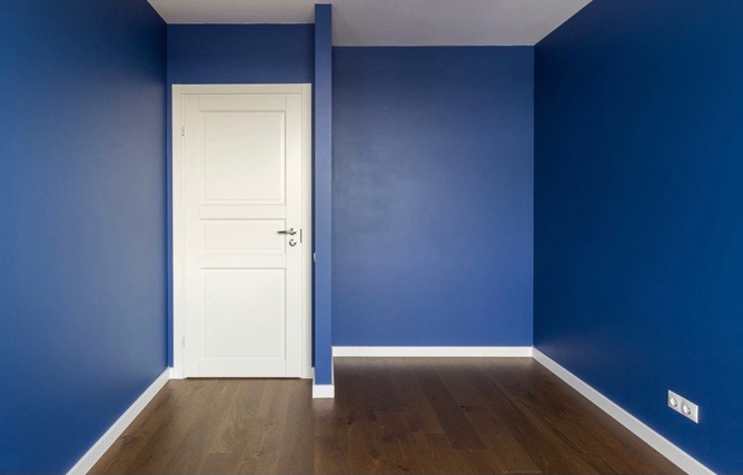 Pomalowane na granatowo wnętrze z drewnianą podłogą i białą stolarką drzwiową oraz listwami przypodłogowymi. Gładka, matowa powierzchnia ścian. Widoczne gniazdko elektryczne.