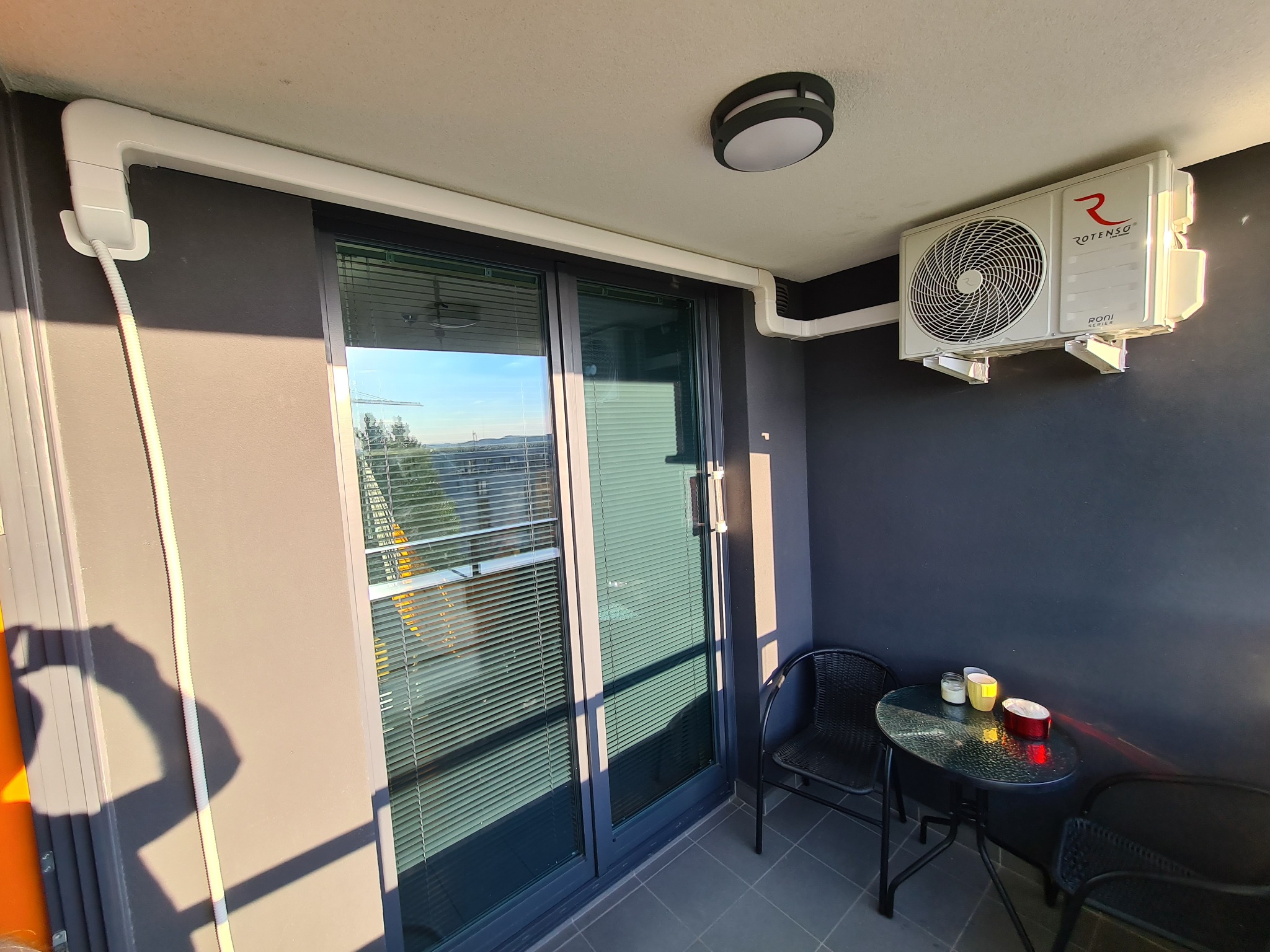 Zewnętrzna jednostka klimatyzatora Rotenso zamontowana na szarej ścianie balkonu, z widocznymi rurami odprowadzającymi i lampą sufitową. Na balkonie stolik z krzesłami.