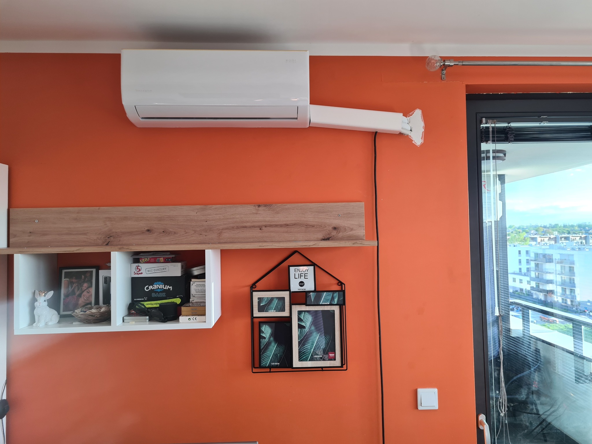 Biały klimatyzator ścienny zamontowany w pomarańczowym pokoju, widoczne rury doprowadzające, półka z dekoracjami i widok z okna na miasto.