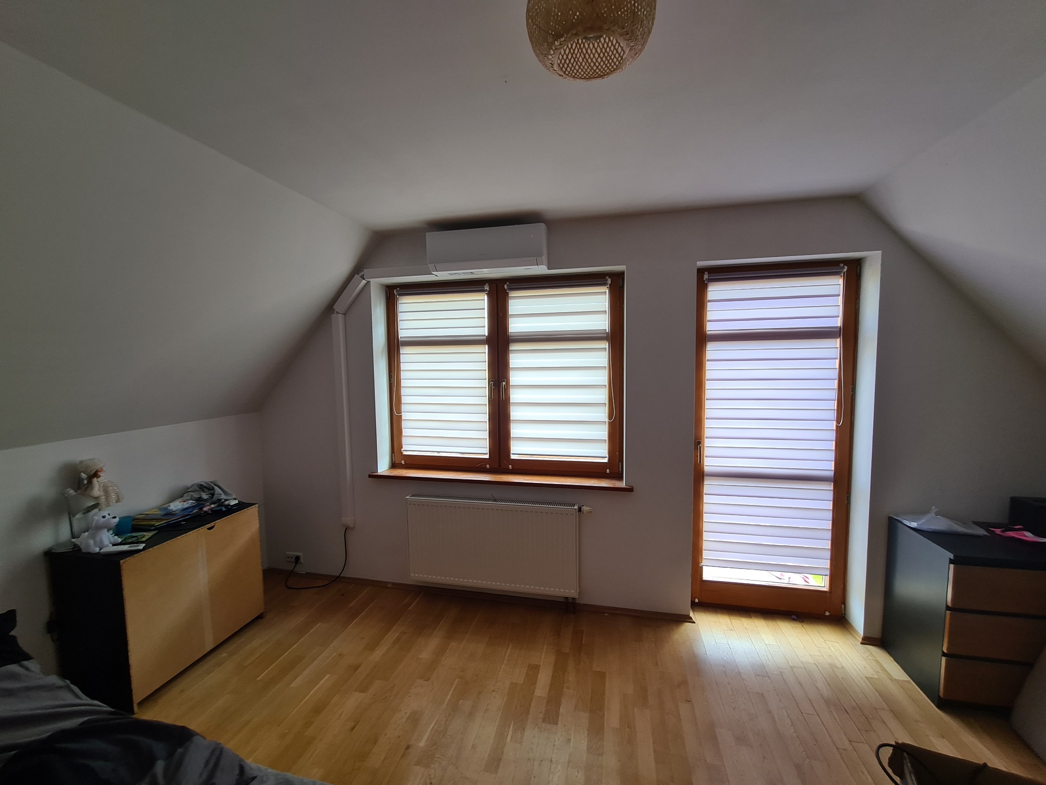 Klimatyzator zamontowany na ścianie w pokoju na poddaszu, widok na okno z żaluzjami i drewnianą ramą, pod oknem grzejnik.