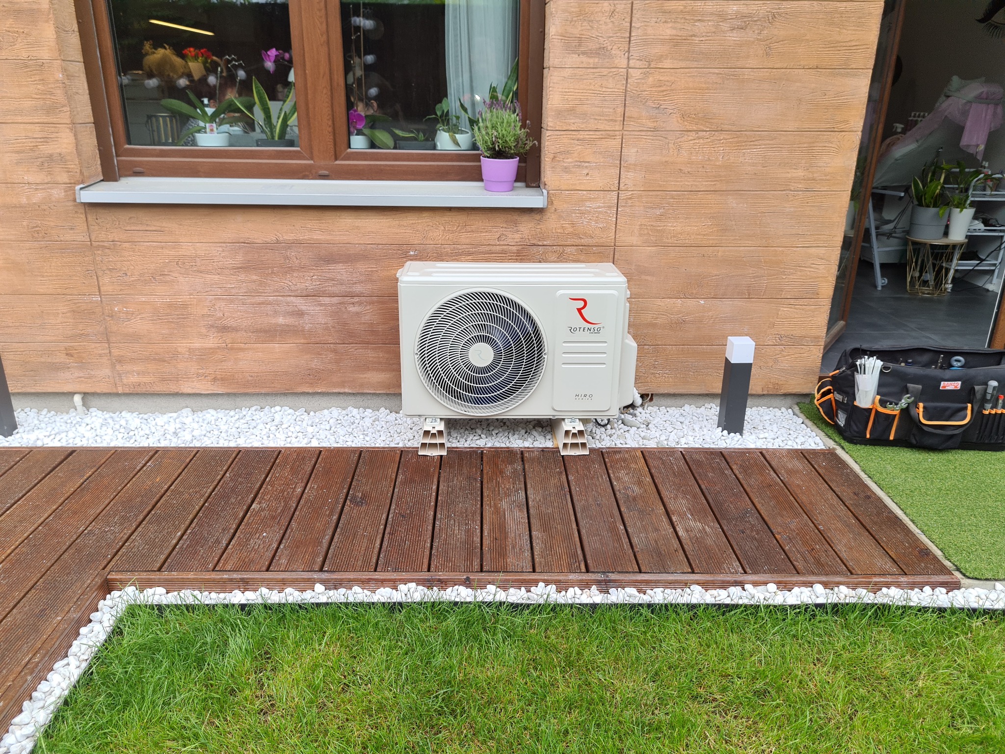 Zewnętrzna jednostka klimatyzacji Rotenso Hiro zamontowana na drewnianym tarasie z białymi kamieniami, widoczny fragment ogrodu i narzędzia w torbie obok.