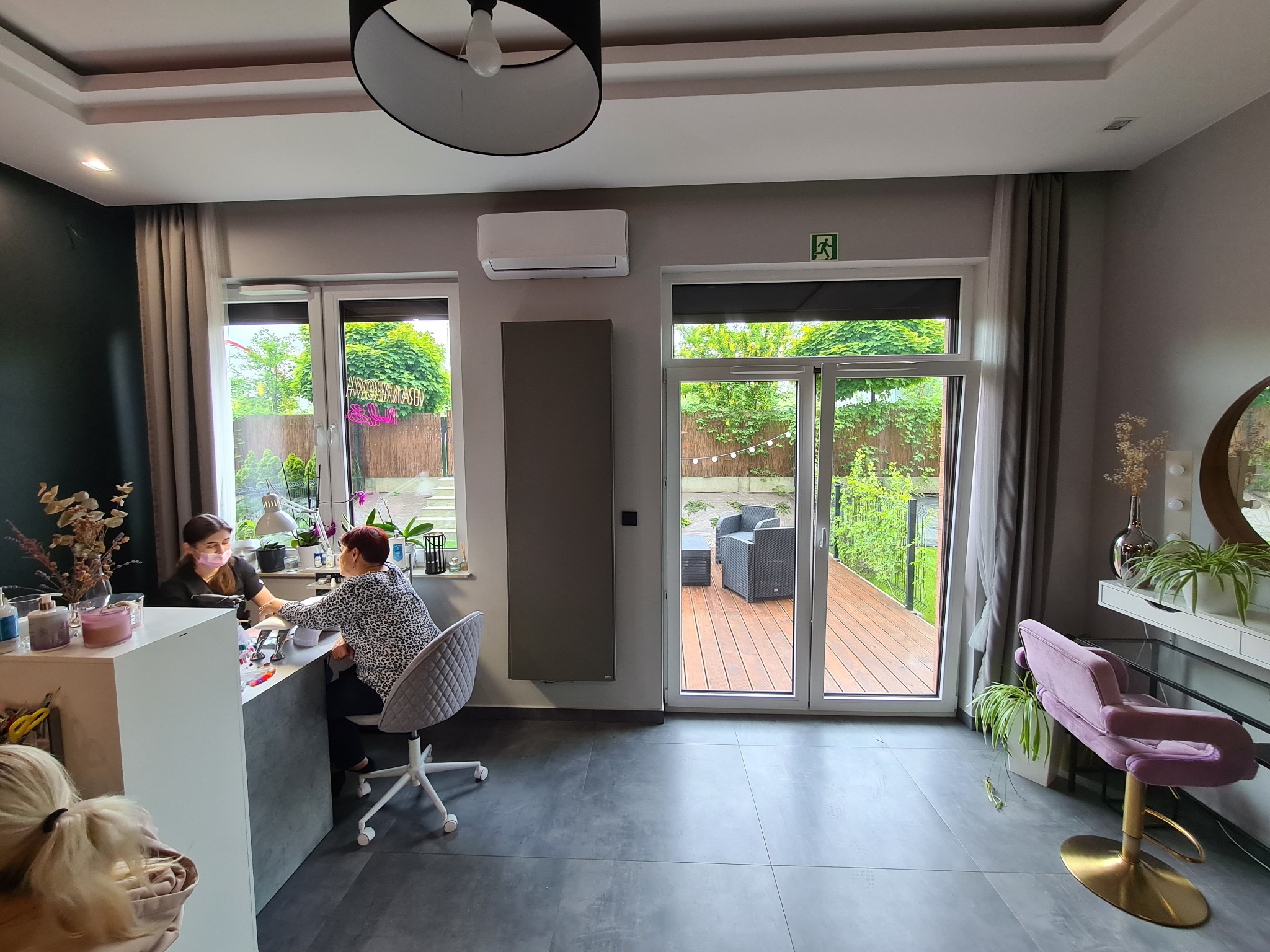 Wnętrze salonu kosmetycznego z widocznym urządzeniem klimatyzacyjnym na ścianie, podczas wykonywania manicure przez stylistkę w maseczce, z widokiem na ogród przez przeszklone drzwi.