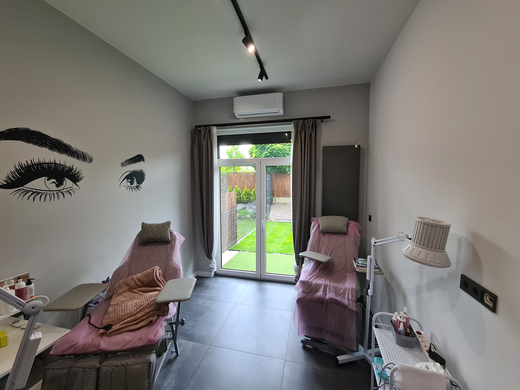 Wnętrze gabinetu kosmetycznego z dwoma fotelami zabiegowymi, klimatyzatorem na ścianie nad oknem balkonowym i naklejkami z motywem oka na ścianie.