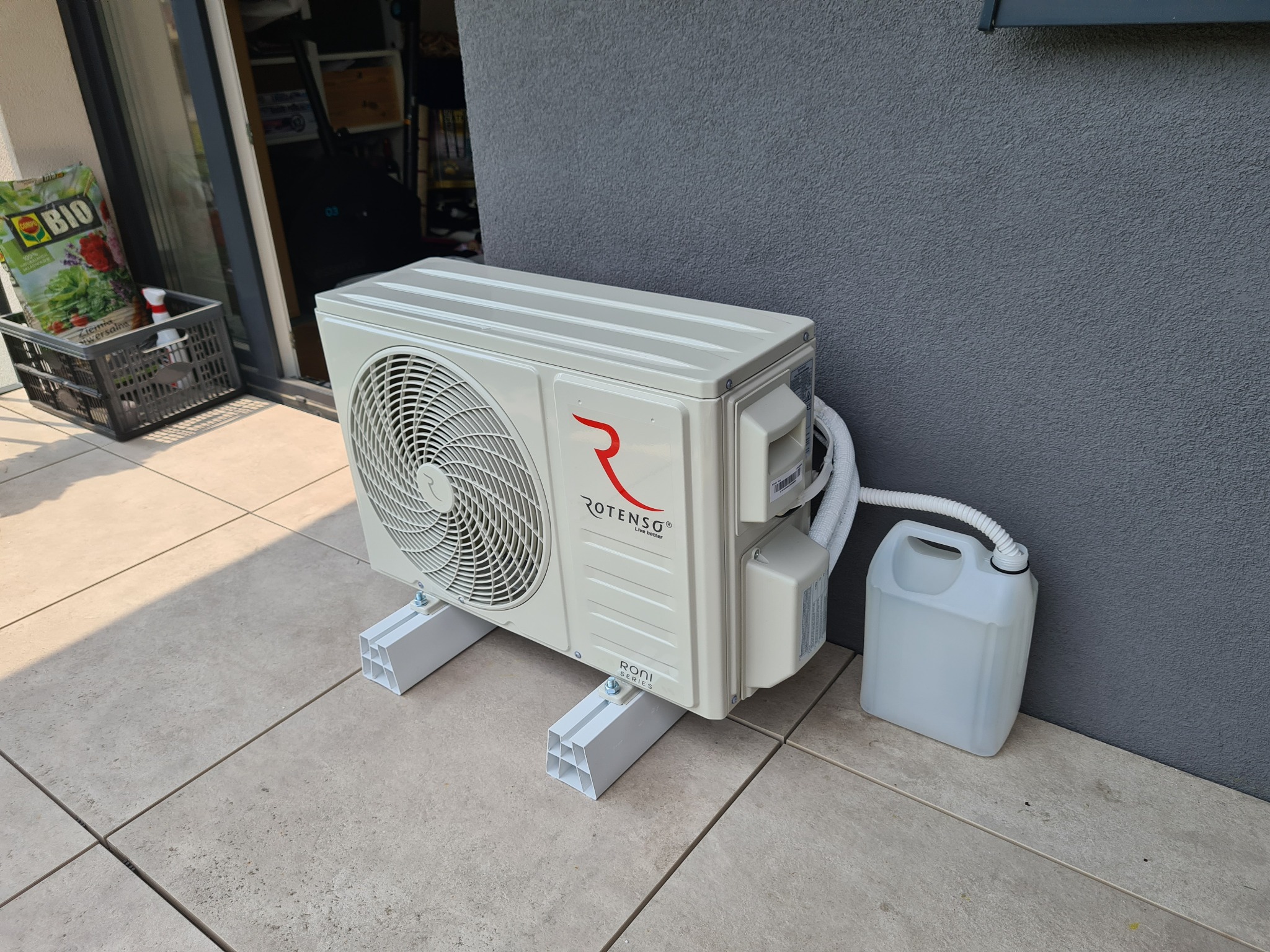 Biała jednostka zewnętrzna klimatyzatora Rotenso zamontowana na białych profilach na jasnych płytkach tarasowych, obok szara ściana i biały kanister na skropliny.