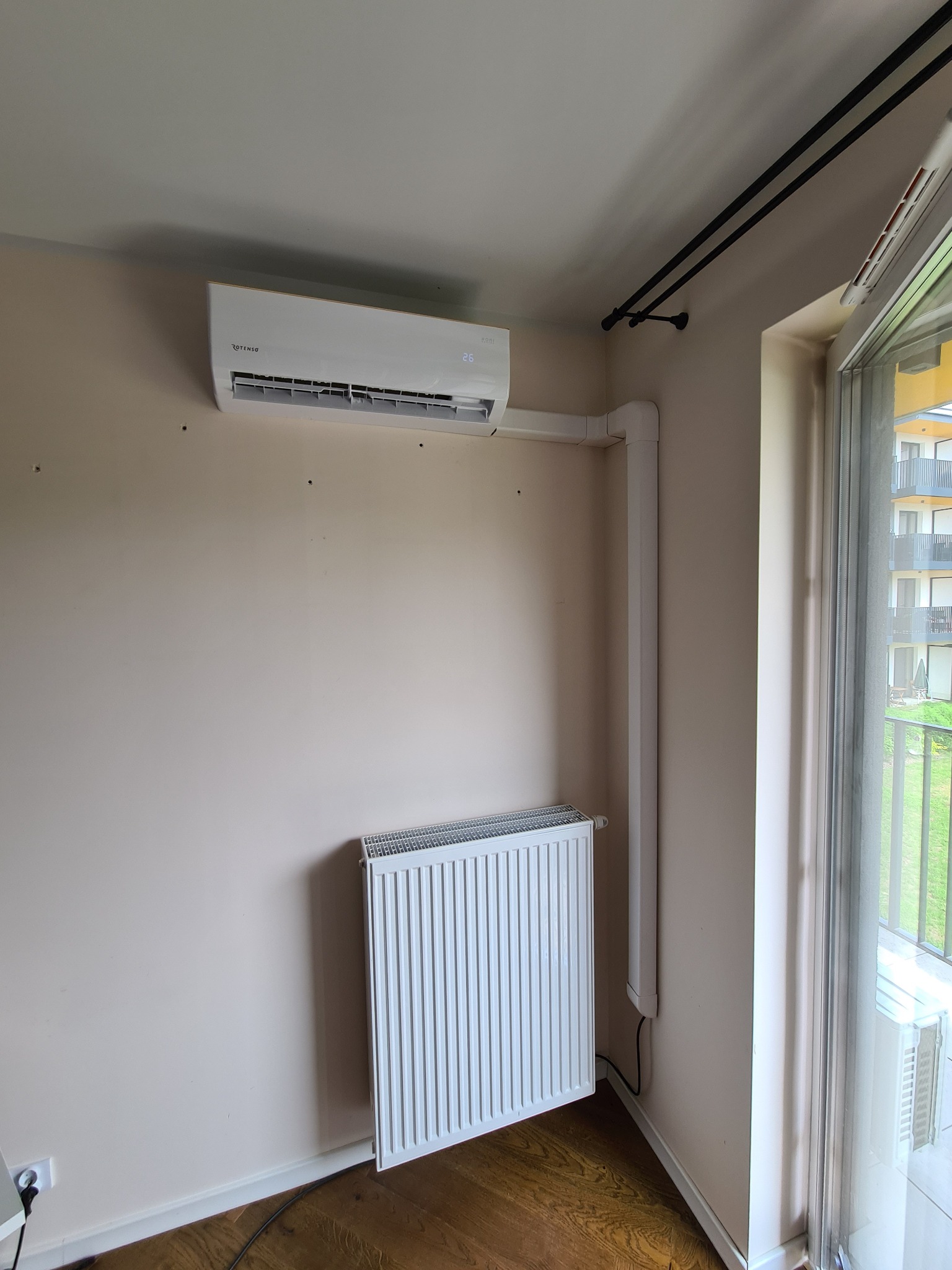 Biały klimatyzator ścienny marki Rotenso zamontowany nad grzejnikiem w pokoju z drewnianą podłogą, widoczne rury odprowadzające skropliny i otwory po wierceniu w ścianie.