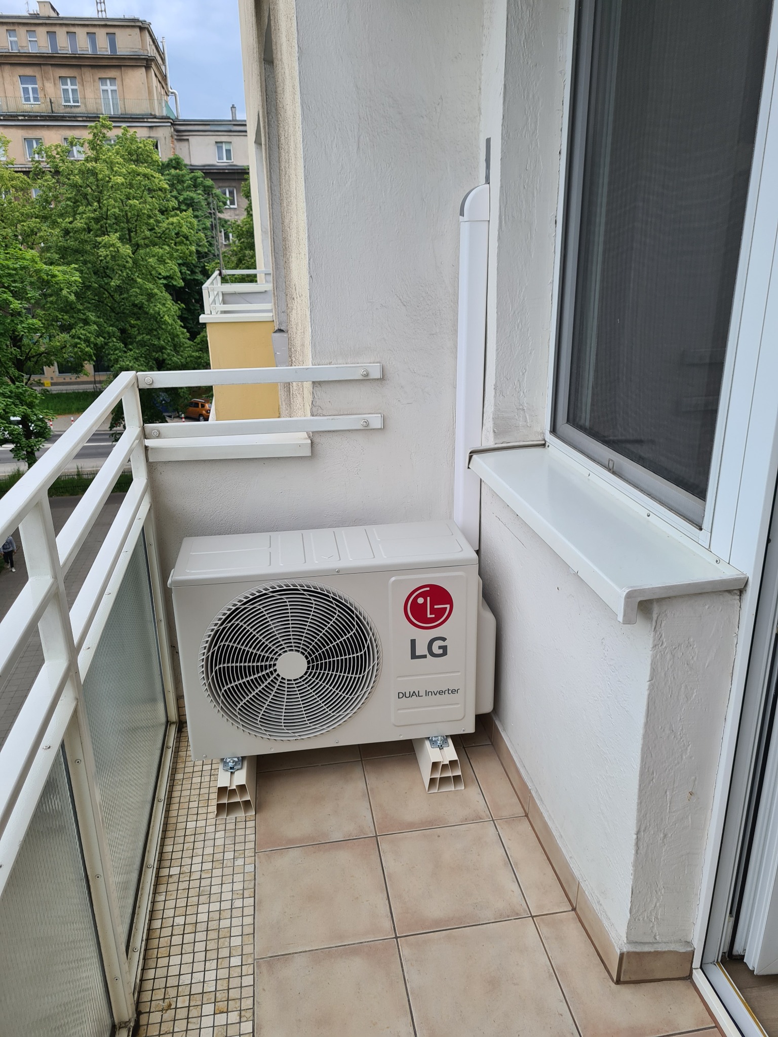 Biała jednostka zewnętrzna klimatyzacji LG Dual Inverter zamontowana na balkonie w bloku mieszkalnym, widok z boku, jasne płytki podłogowe.