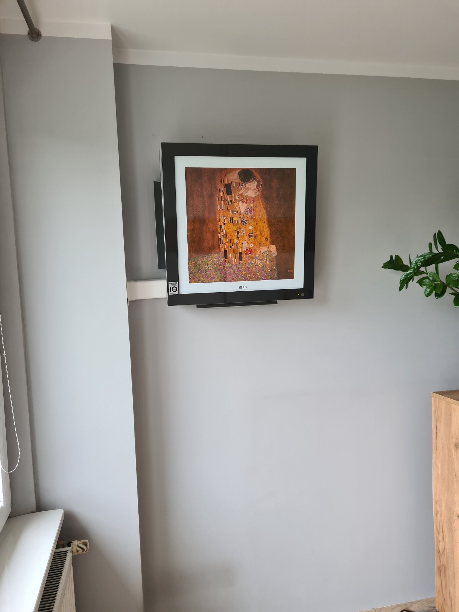Telewizor LG zamontowany na szarej ścianie w salonie, wyświetlający obraz 'Pocałunek' Gustava Klimta, obok roślina doniczkowa.
