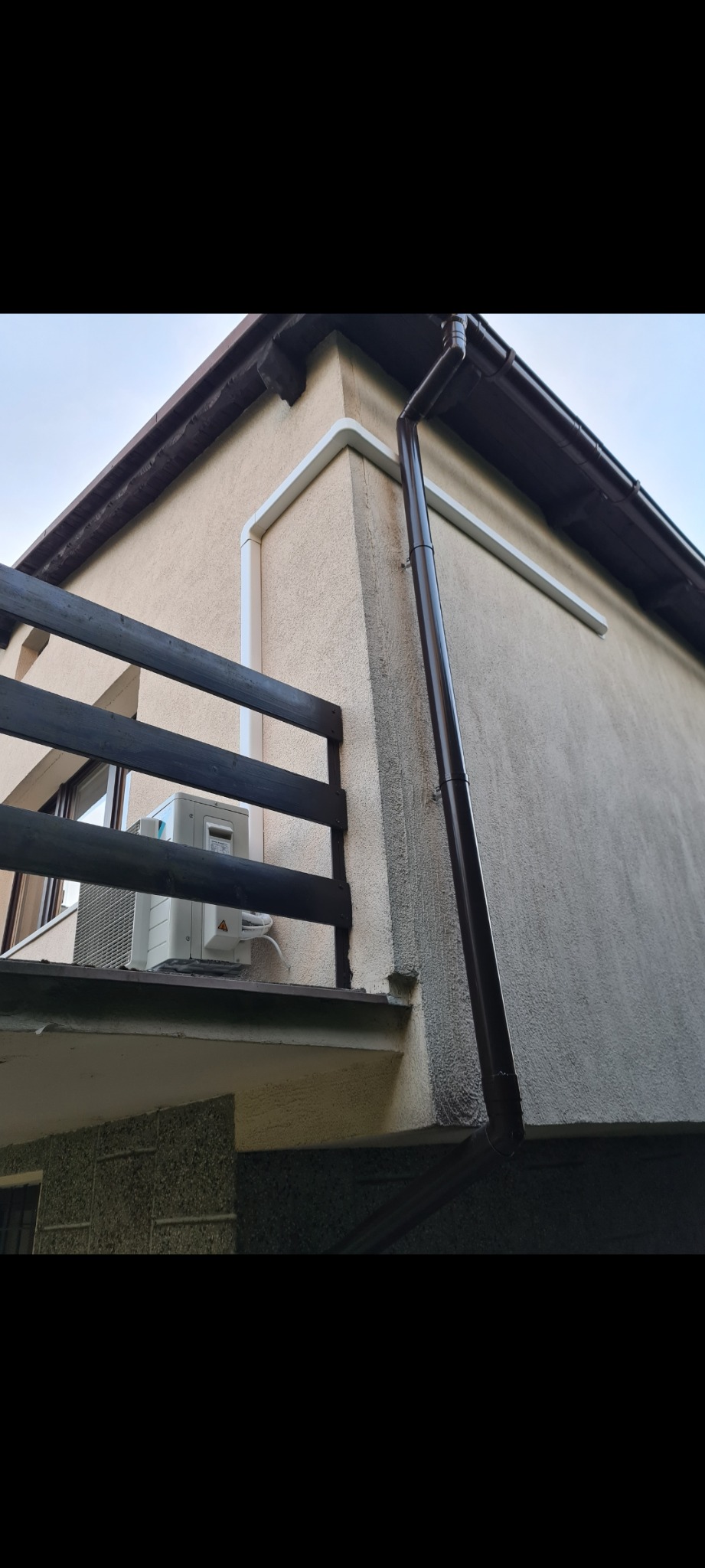 Zewnętrzna jednostka klimatyzacji zamontowana na balkonie budynku mieszkalnego, widoczna z dołu, z brązową rynną i ciemnym drewnianym balustradą.