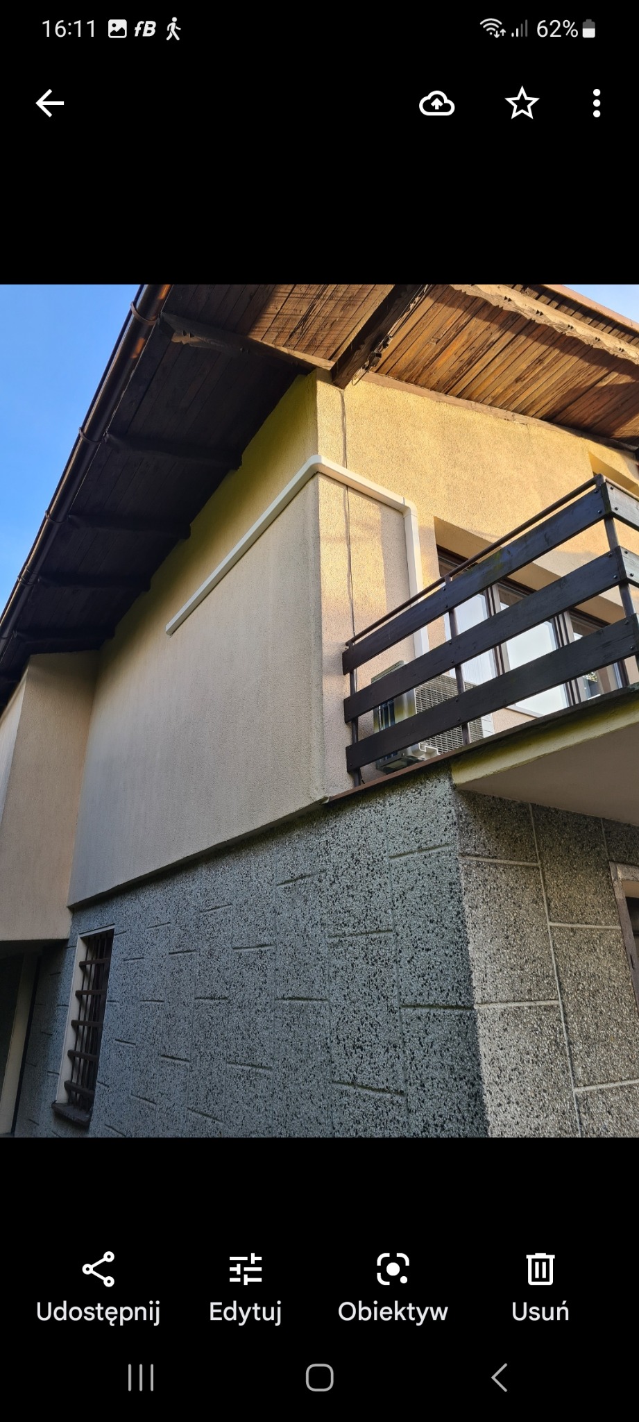 Zewnętrzna jednostka klimatyzacji zamontowana na balkonie domu, widoczna z dołu, z drewnianą balustradą i elewacją budynku w tle.