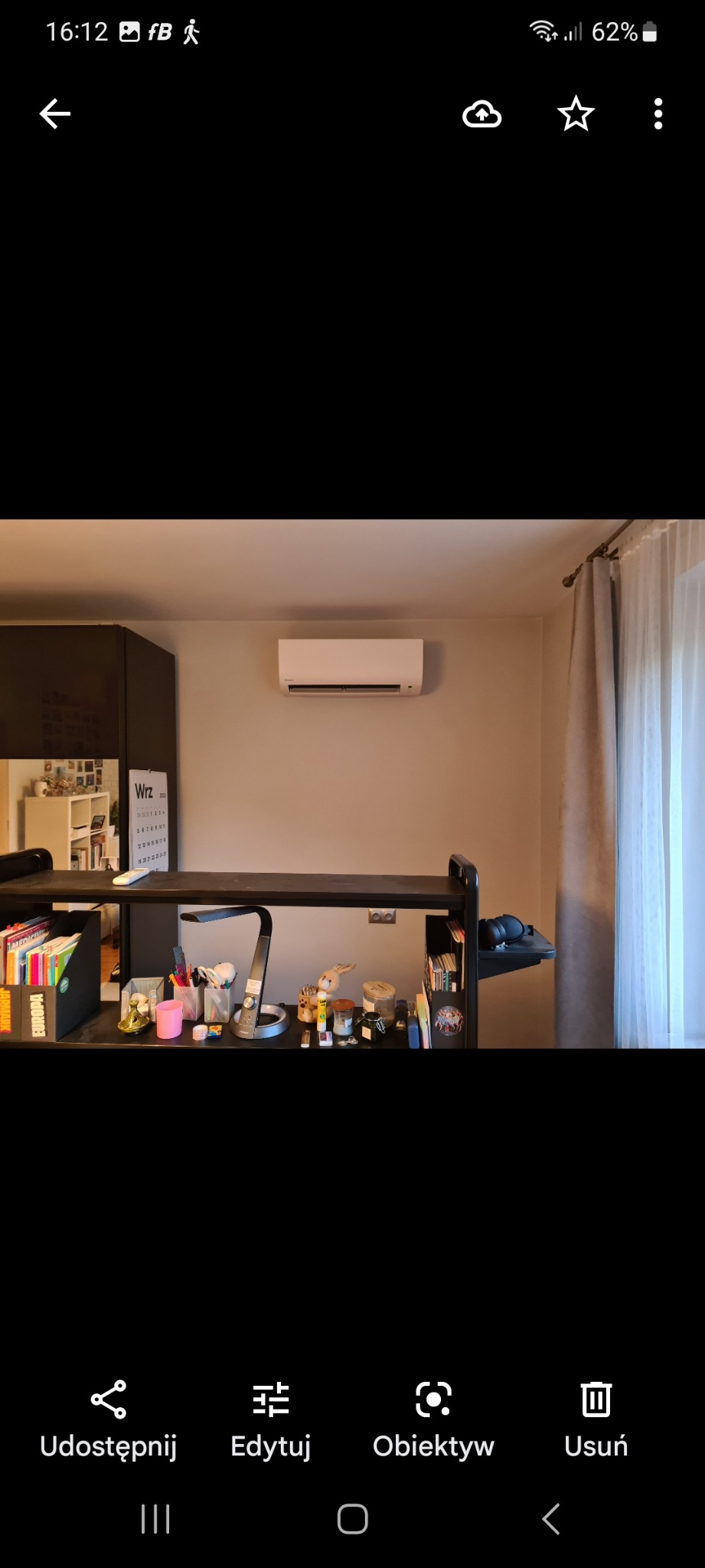 Biały klimatyzator ścienny zamontowany w pokoju nad biurkiem z lampką i akcesoriami, widok na okno z zasłoną.