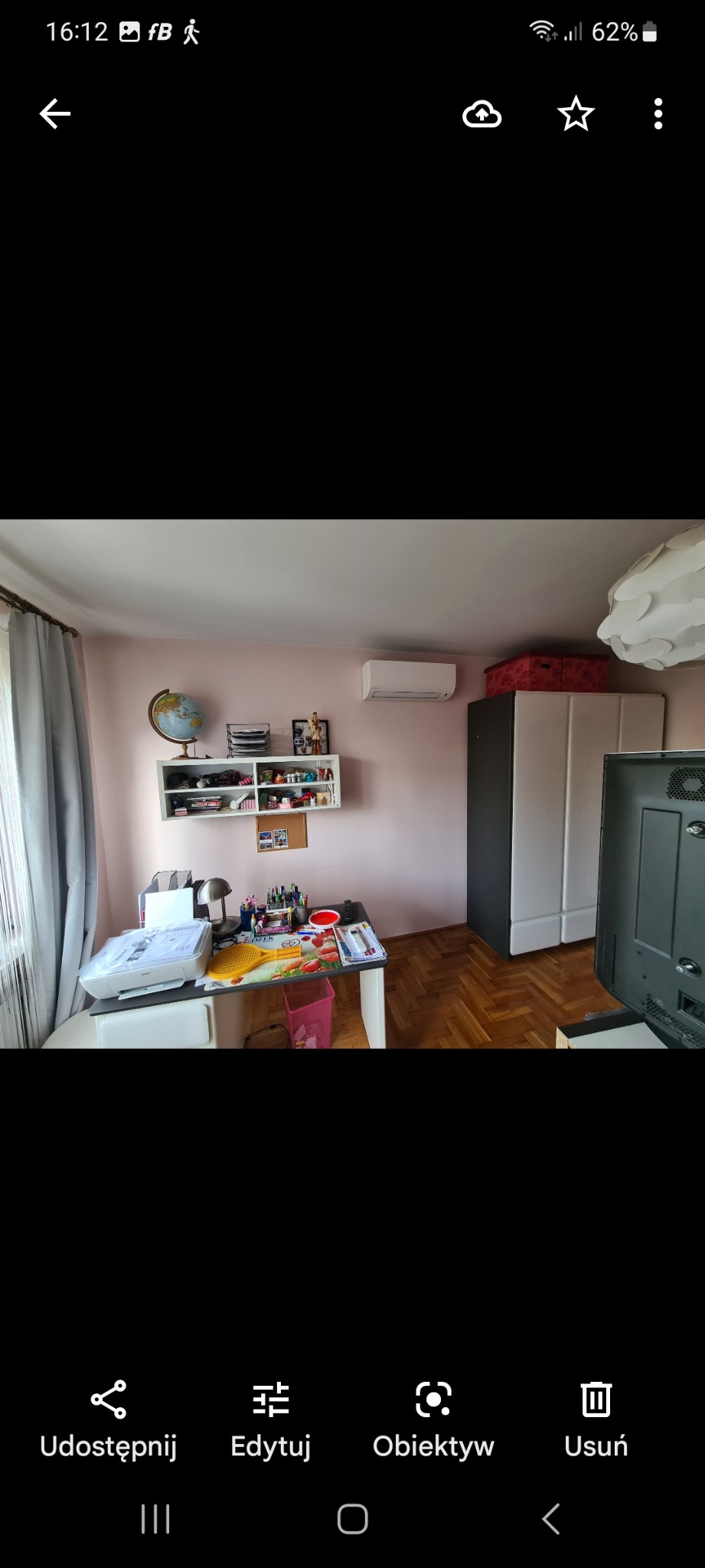 Biała jednostka klimatyzacyjna zamontowana na ścianie w pokoju nad półką z globusem i dekoracjami, widok na biurko z przyborami biurowymi i drukarką.