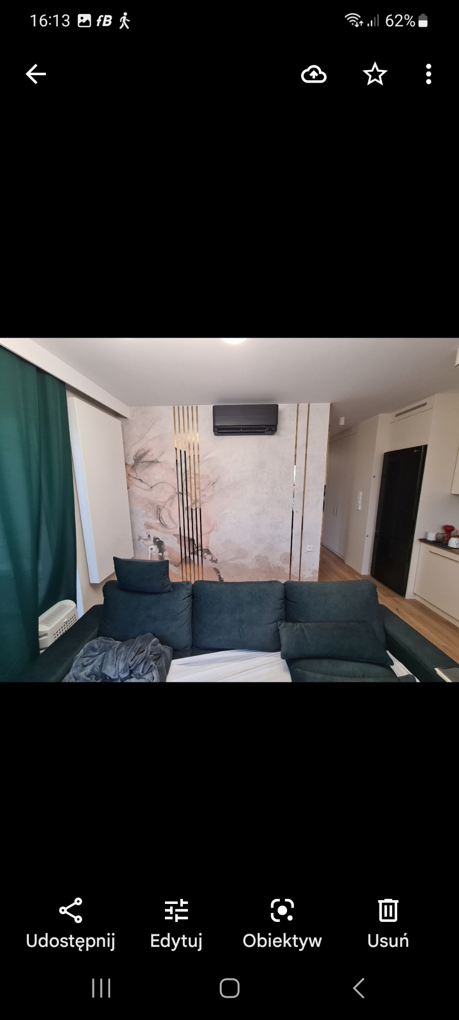 Czarny klimatyzator ścienny zamontowany w salonie nad zieloną kanapą, widok na ścianę z dekoracyjnymi złotymi paskami i wzorem w odcieniach różu.