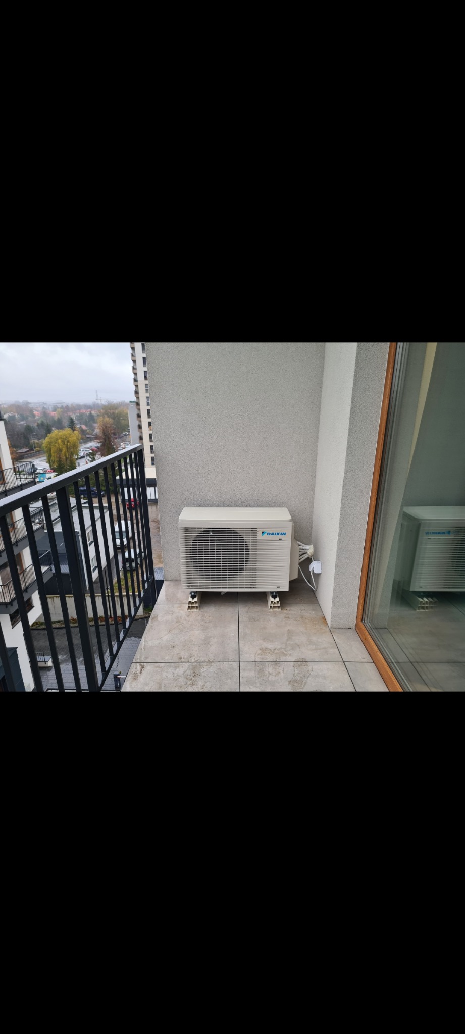 Biała jednostka zewnętrzna klimatyzacji Daikin zamontowana na balkonie bloku mieszkalnego, widok na szarą pogodę za barierką.