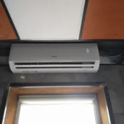 Biała jednostka klimatyzacyjna marki Gree zamontowana na ścianie nad oknem, widoczny wyświetlacz temperatury ustawionej na 22 stopnie.