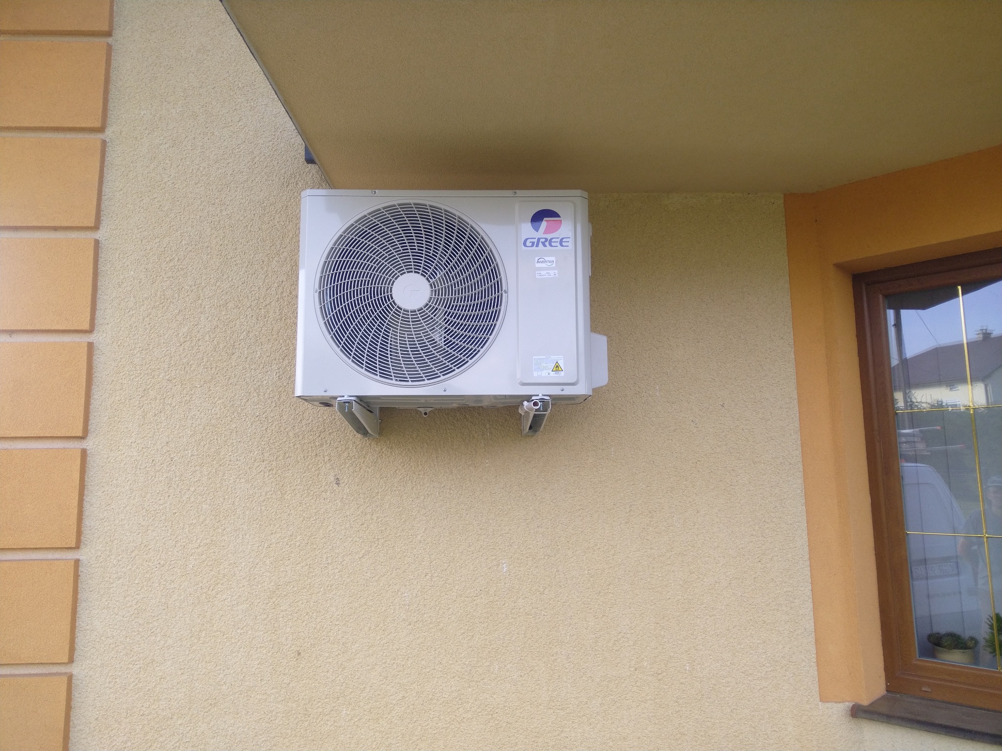 Zewnętrzna jednostka klimatyzacji marki Gree zamontowana na beżowej, teksturowanej ścianie budynku mieszkalnego, widoczna z boku okna z brązową ramą.