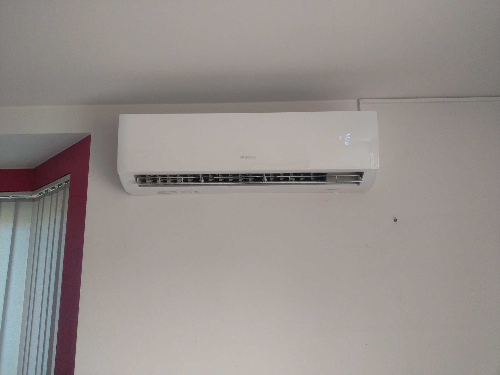 Biały klimatyzator marki Gree zamontowany na ścianie, z wyświetlaczem wskazującym 16 stopni Celsjusza, obok okna z żaluzjami w bordowej ramie.