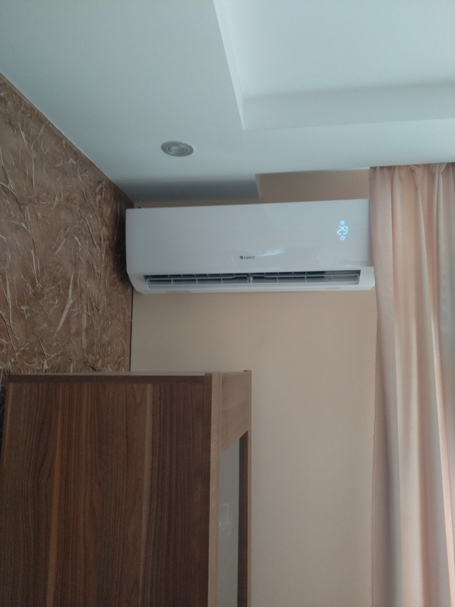 Biała klimatyzacja marki Gree zamontowana w pokoju, widoczny wyświetlacz temperatury, obok zasłona i fragment szafy.