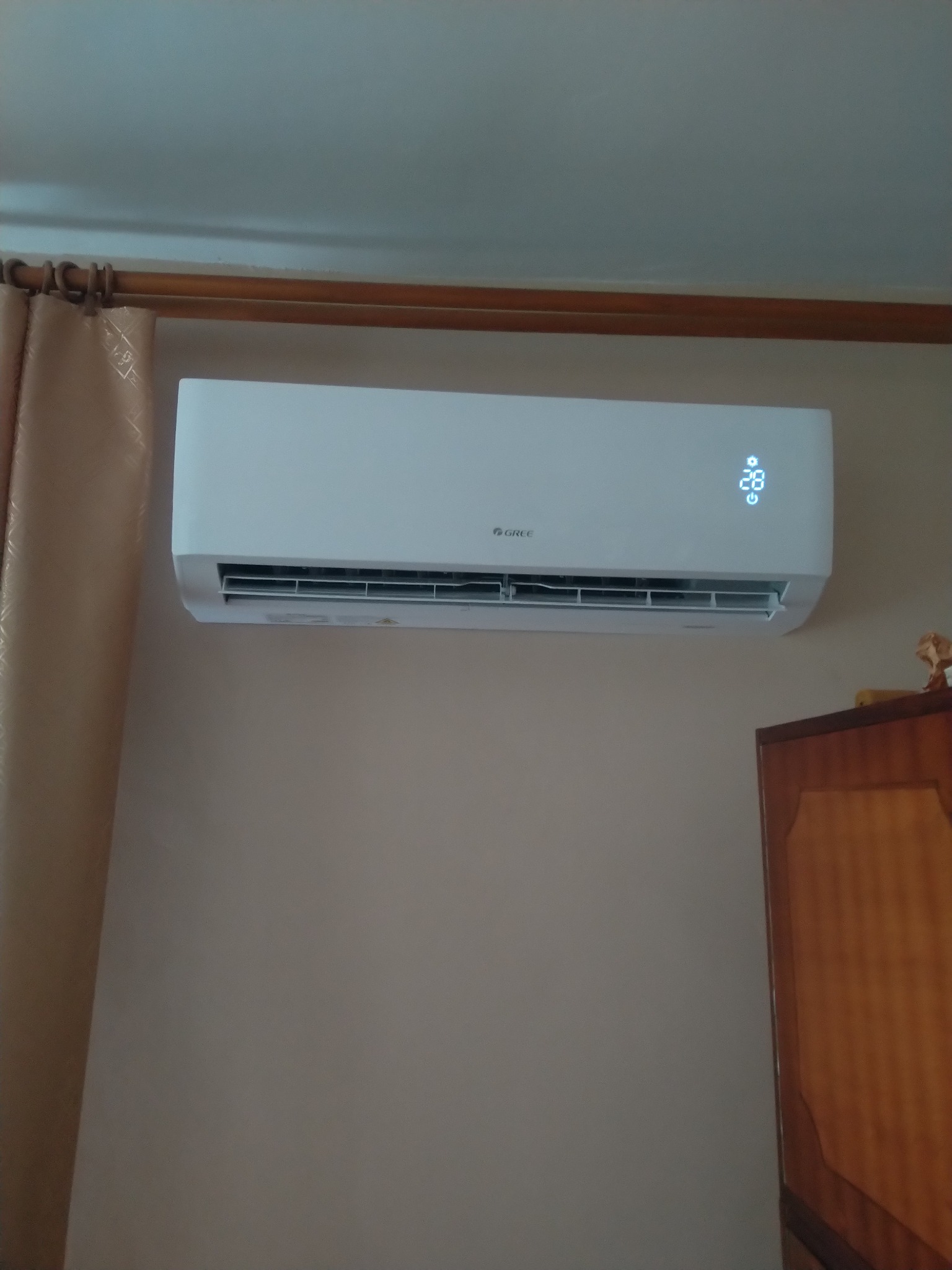 Biała jednostka klimatyzacyjna marki Gree zamontowana na ścianie w pomieszczeniu mieszkalnym, wyświetlająca temperaturę 28 stopni Celsjusza, widoczny fragment zasłony i szafki.