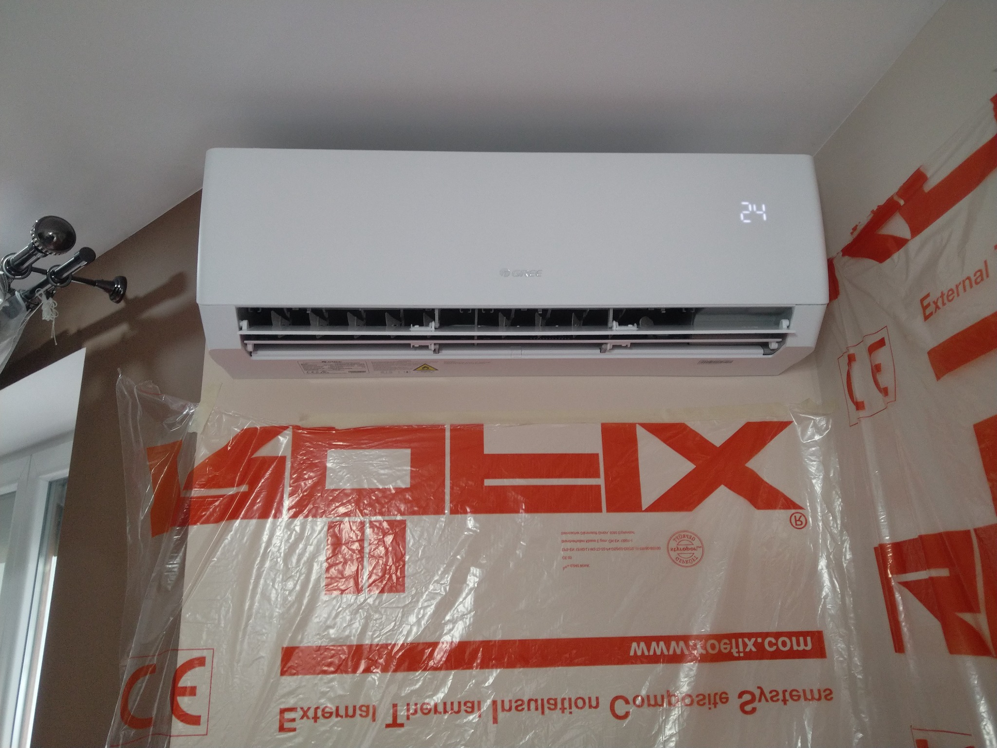 Biała jednostka klimatyzacyjna Gree zamontowana na ścianie, zabezpieczonej folią ochronną z nadrukiem w kolorze pomarańczowym. Wyświetlacz pokazuje temperaturę 24 stopnie.