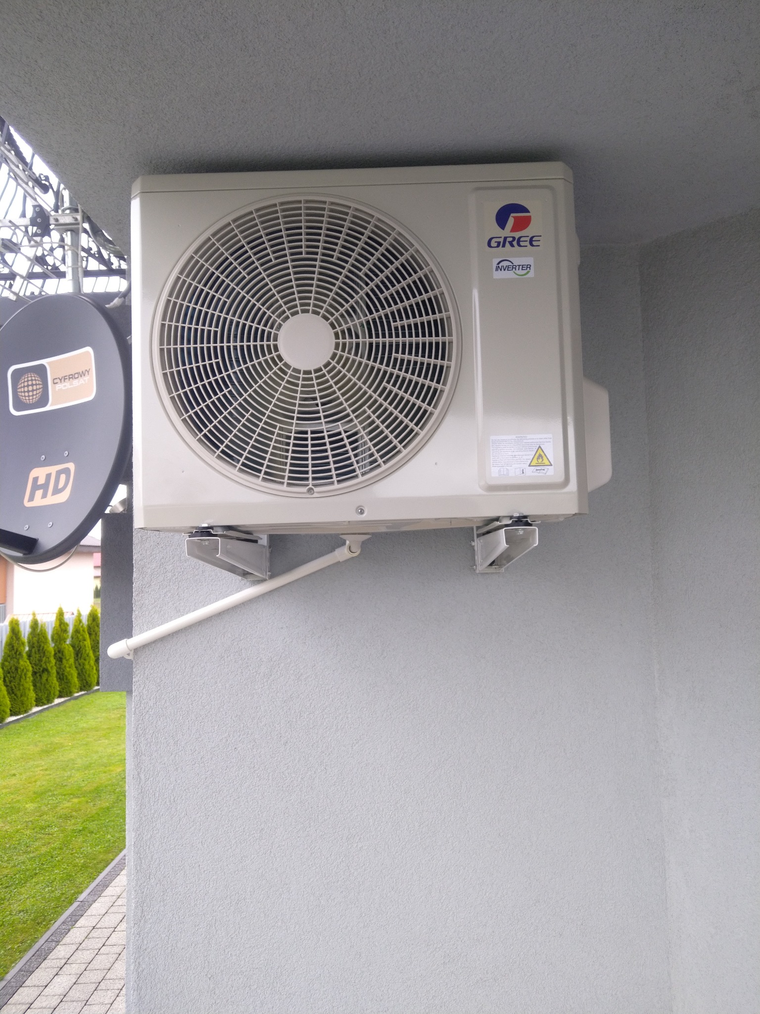 Zewnętrzna jednostka klimatyzacji marki Gree zamontowana na szarej ścianie budynku, obok anteny satelitarnej i widoku na zielony trawnik z tujami.