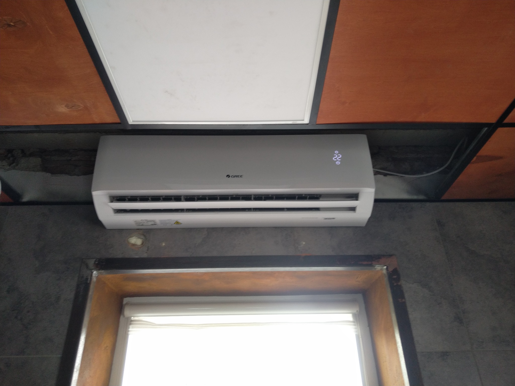Biała jednostka klimatyzacyjna marki Gree zamontowana na ścianie nad oknem, widoczny wyświetlacz temperatury ustawionej na 22 stopnie.
