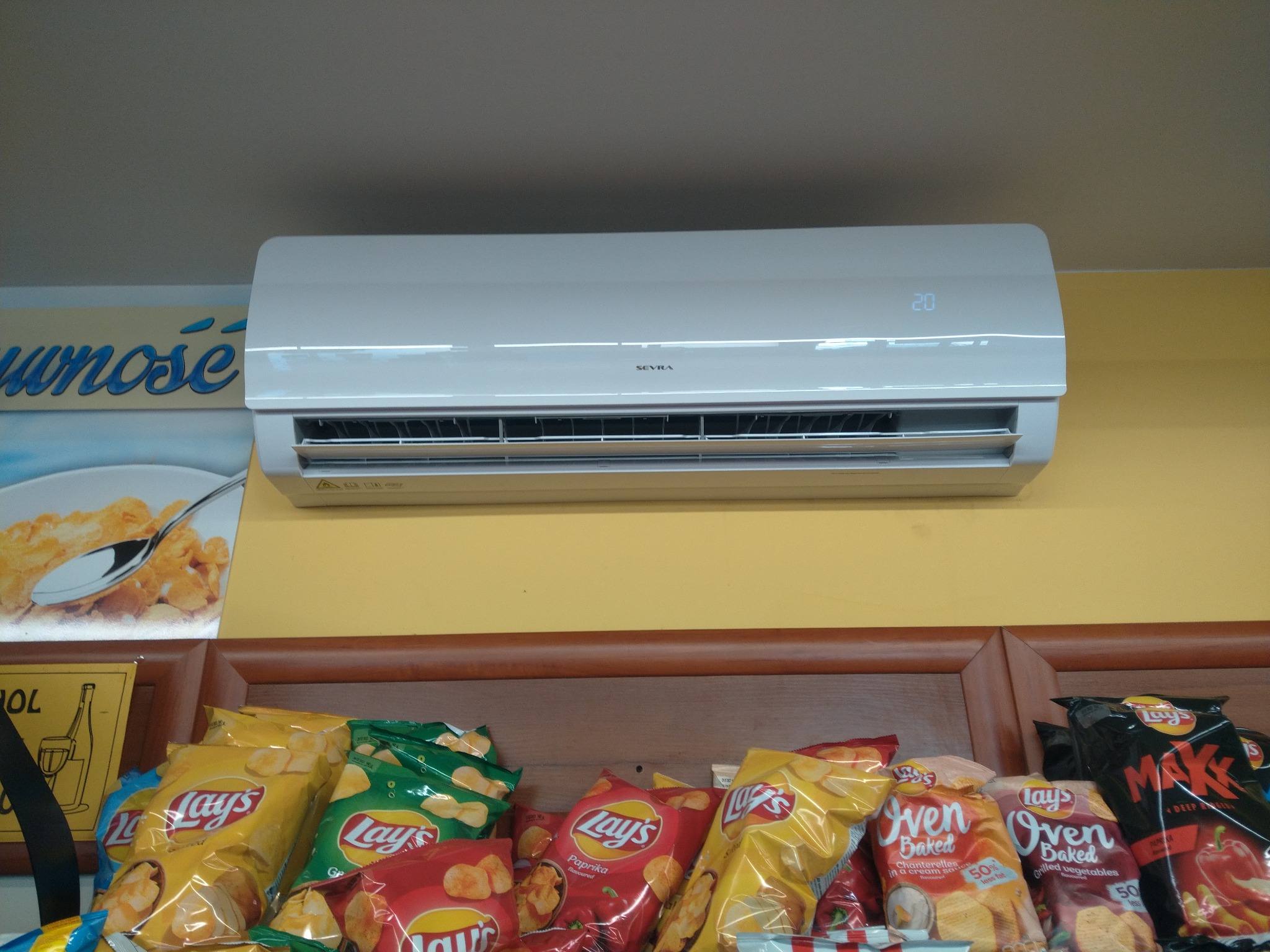 Biały klimatyzator marki Sevra zamontowany nad półką z przekąskami w sklepie, widoczny wyświetlacz z temperaturą 20 stopni.