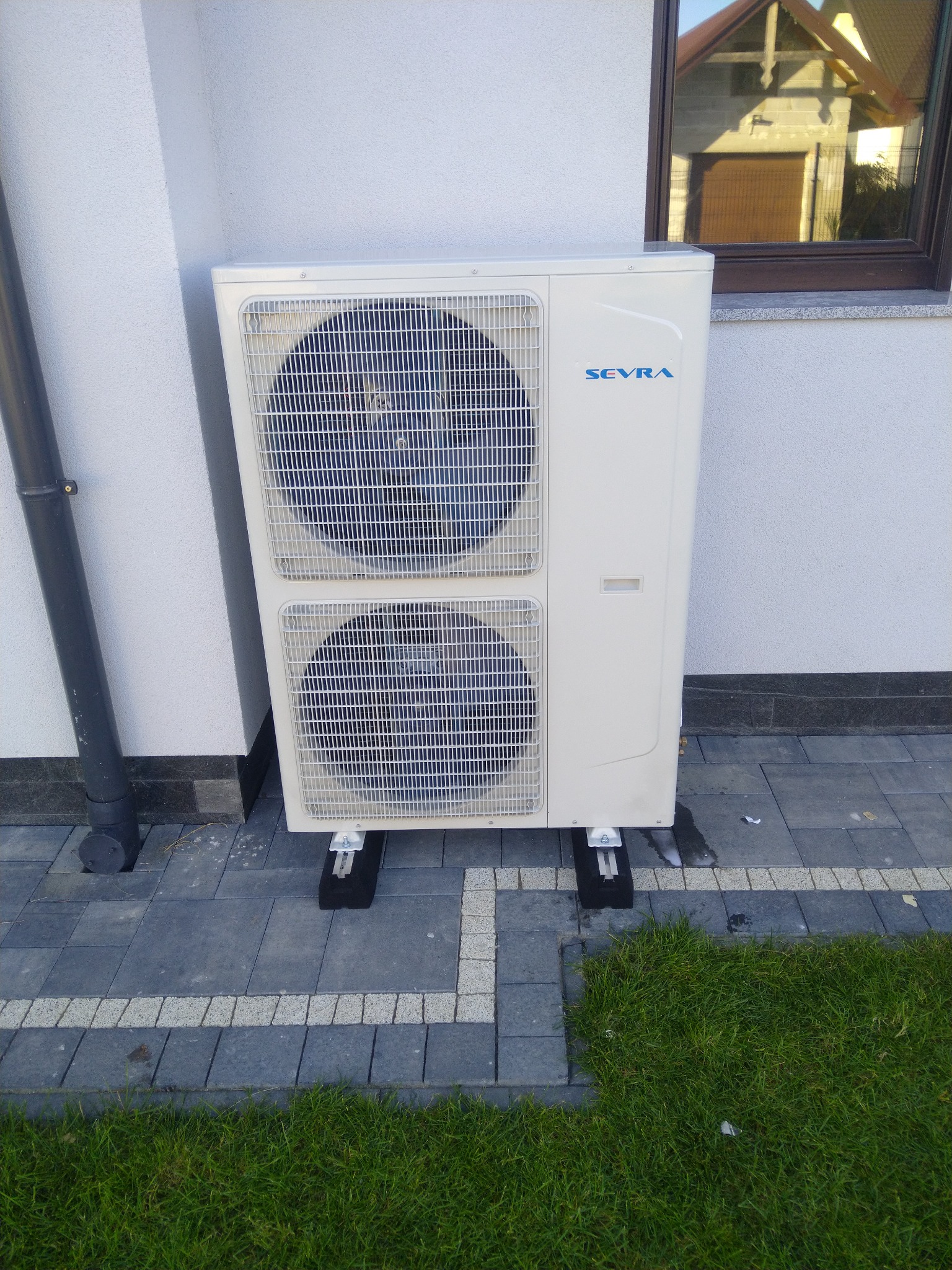 Zewnętrzna jednostka klimatyzacji SEVRA zamontowana na czarnych wspornikach przy ścianie domu z widocznym oknem i fragmentem dachu, obok rynna spustowa, na szarej kostce brukowej i trawniku.