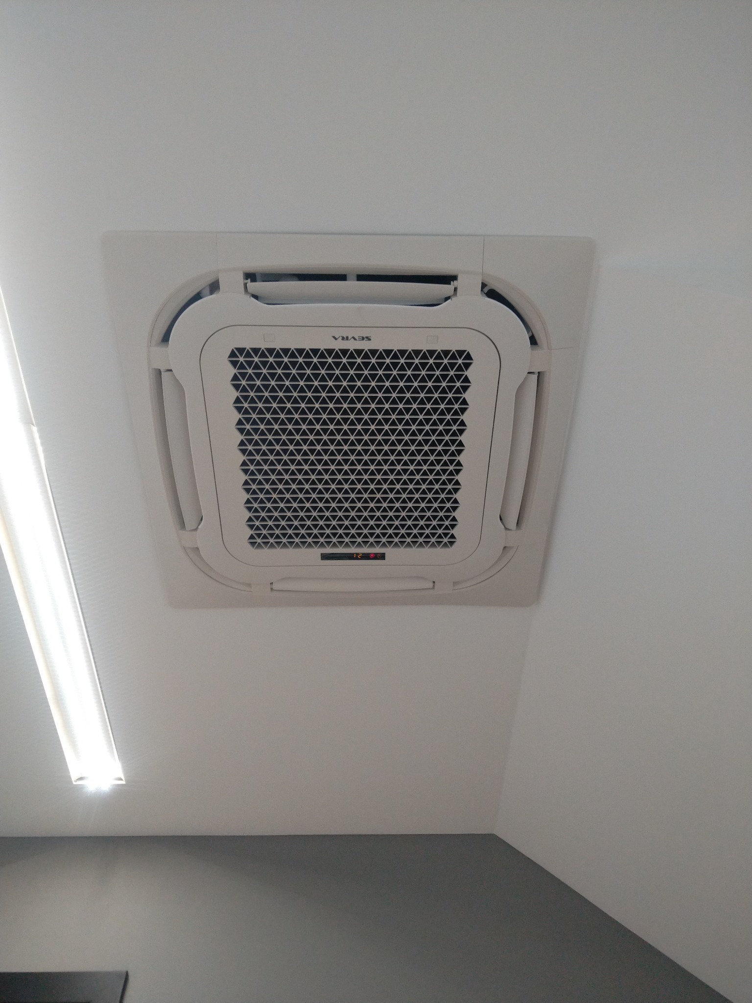 Klimatyzator kasetonowy marki SEVRA zamontowany w suficie biura, widoczny z dołu pod kątem, z wyświetlaczem wskazującym temperaturę 12 stopni Celsjusza.