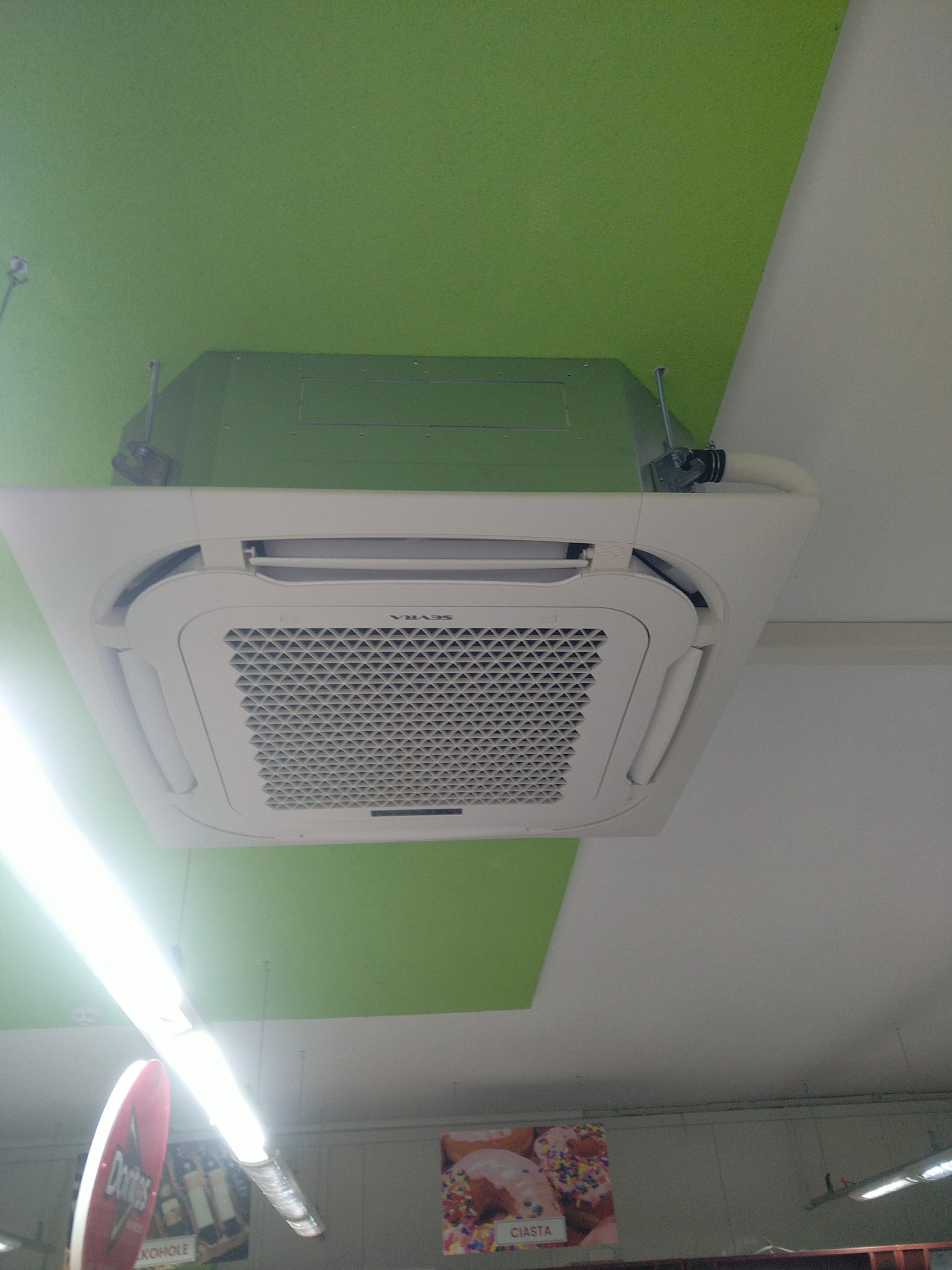 Biała, kasetonowa jednostka klimatyzacji zamontowana na suficie w pomieszczeniu handlowym, widoczne reklamy produktów spożywczych.