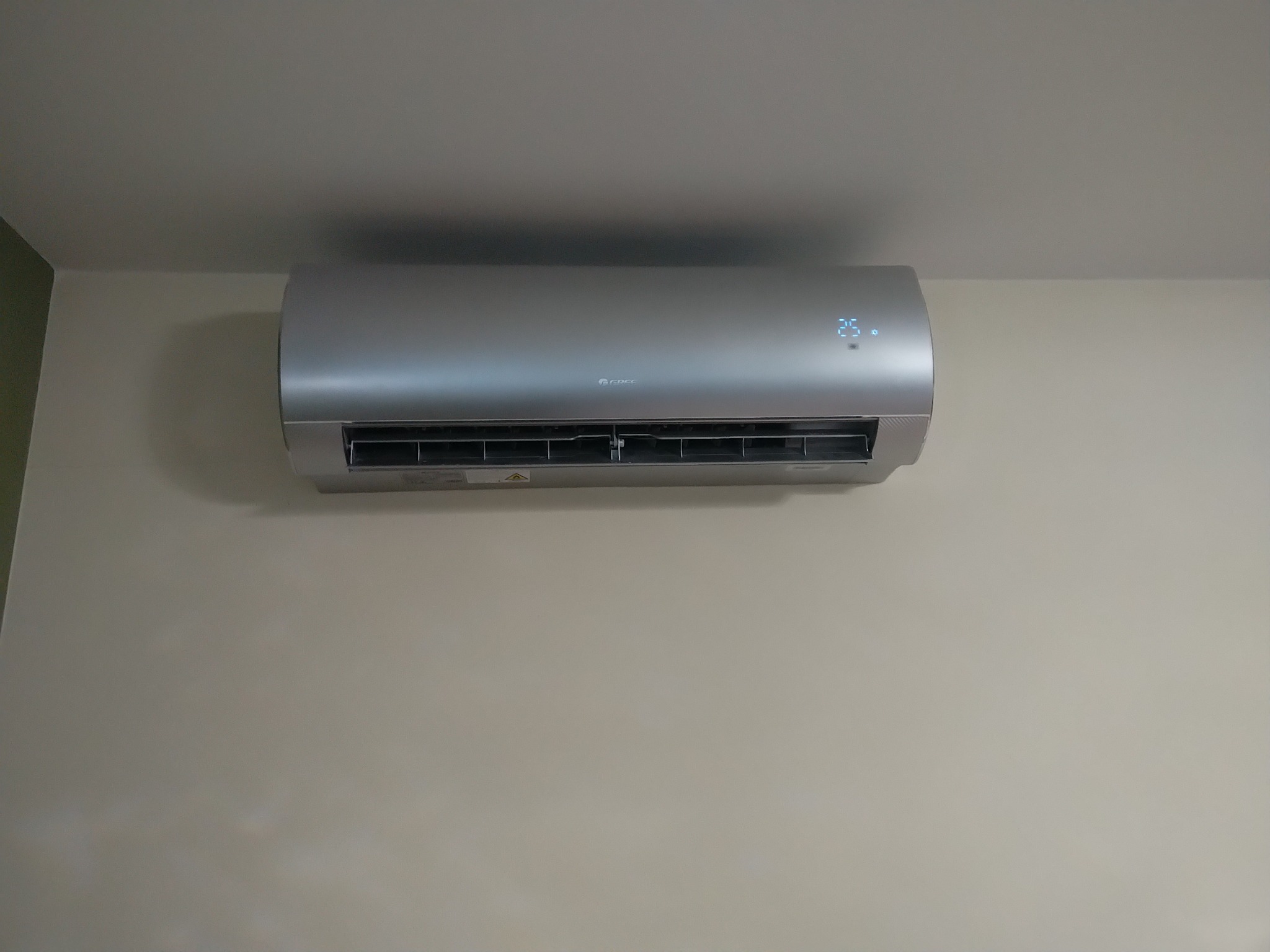 Zamontowany na ścianie, srebrny klimatyzator Gree z wyświetlaną temperaturą 25 stopni Celsjusza, widoczny fragment zielonej ściany po lewej stronie.