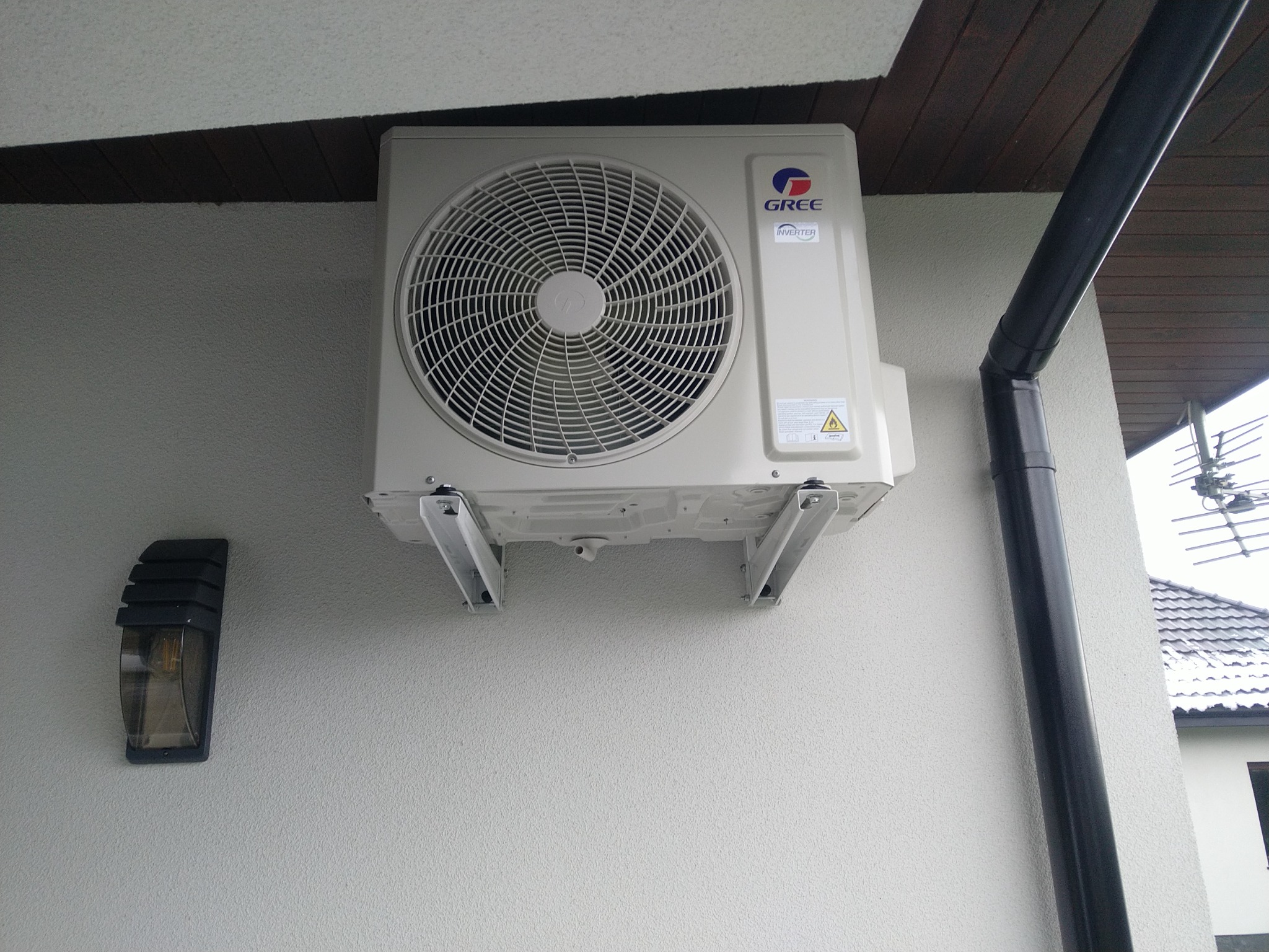 Zewnętrzna jednostka klimatyzacji marki Gree zamontowana na elewacji domu, widoczny fragment rynny i lampa zewnętrzna.