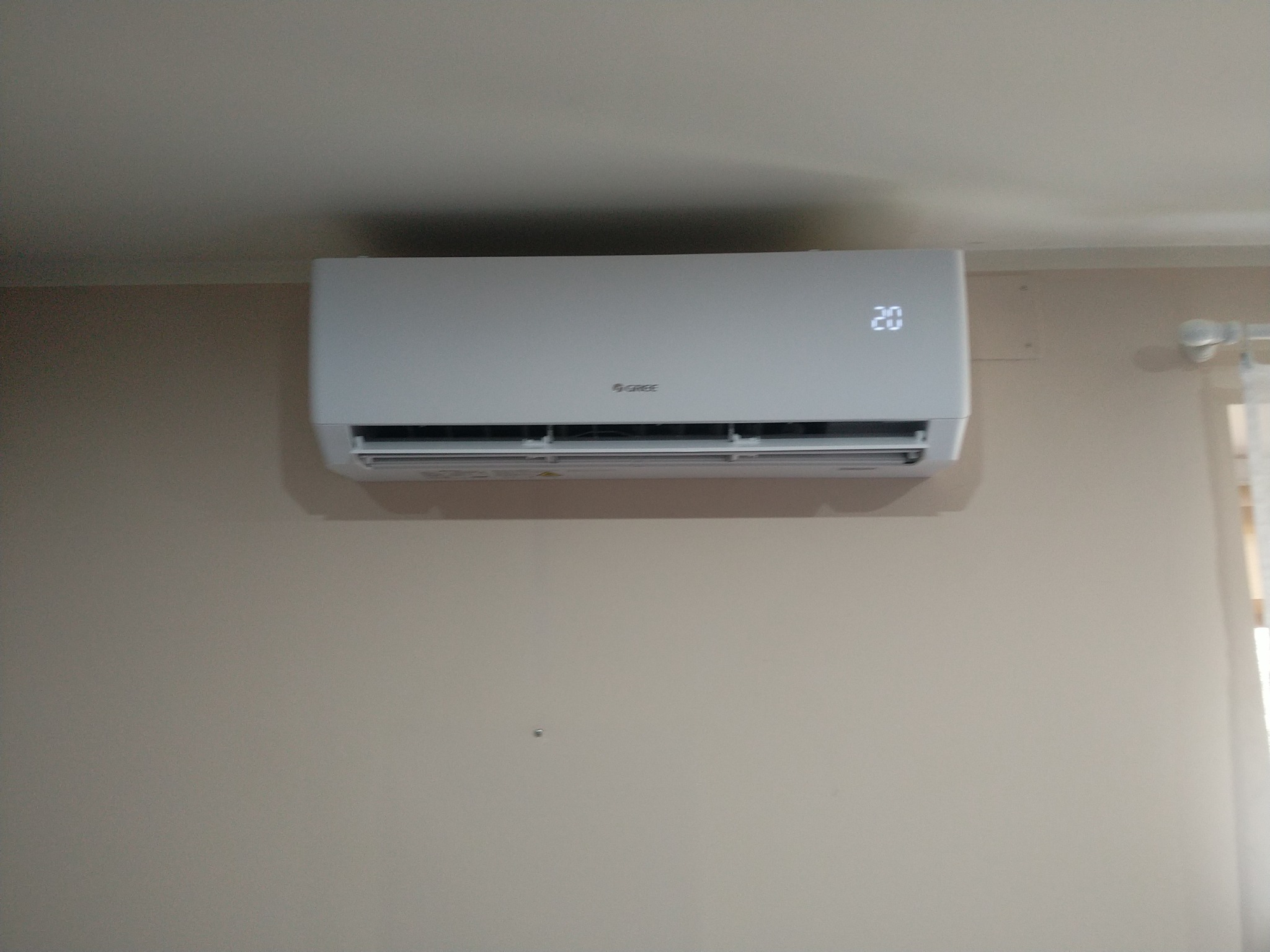 Biały klimatyzator marki Gree zamontowany na beżowej ścianie w pomieszczeniu. Na wyświetlaczu urządzenia widoczna temperatura 20 stopni Celsjusza.