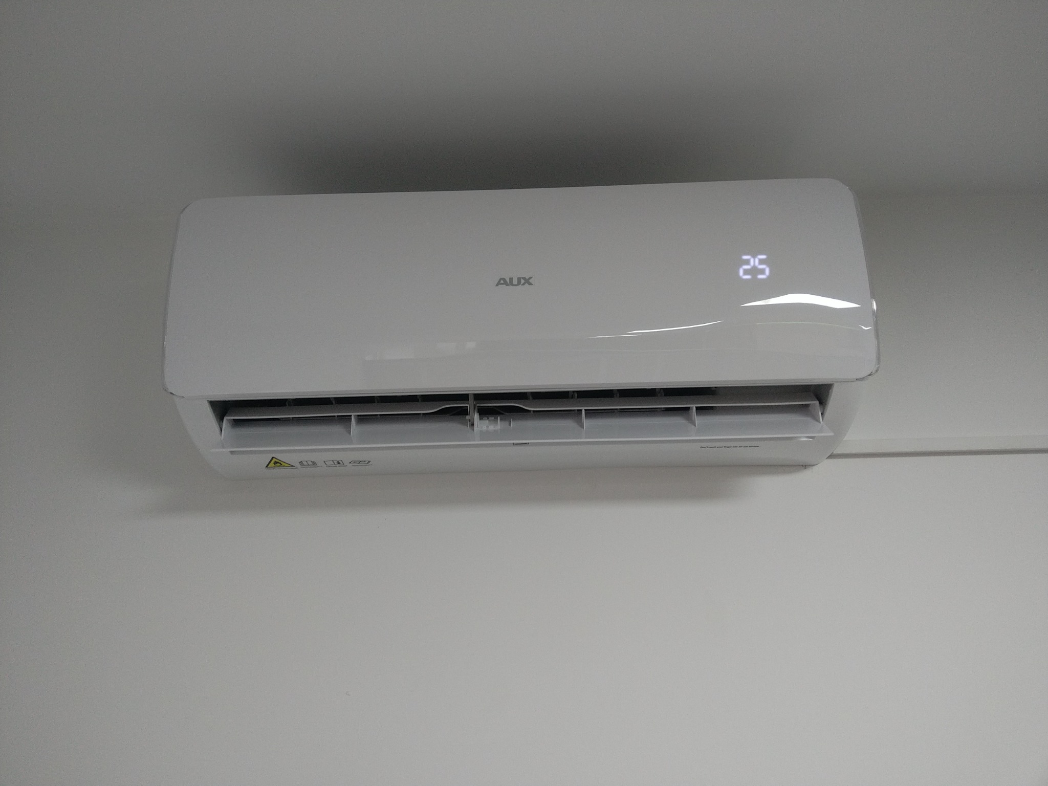 Nowoczesny, biały klimatyzator marki AUX zamontowany na białej ścianie, wyświetlający temperaturę 25 stopni Celsjusza.