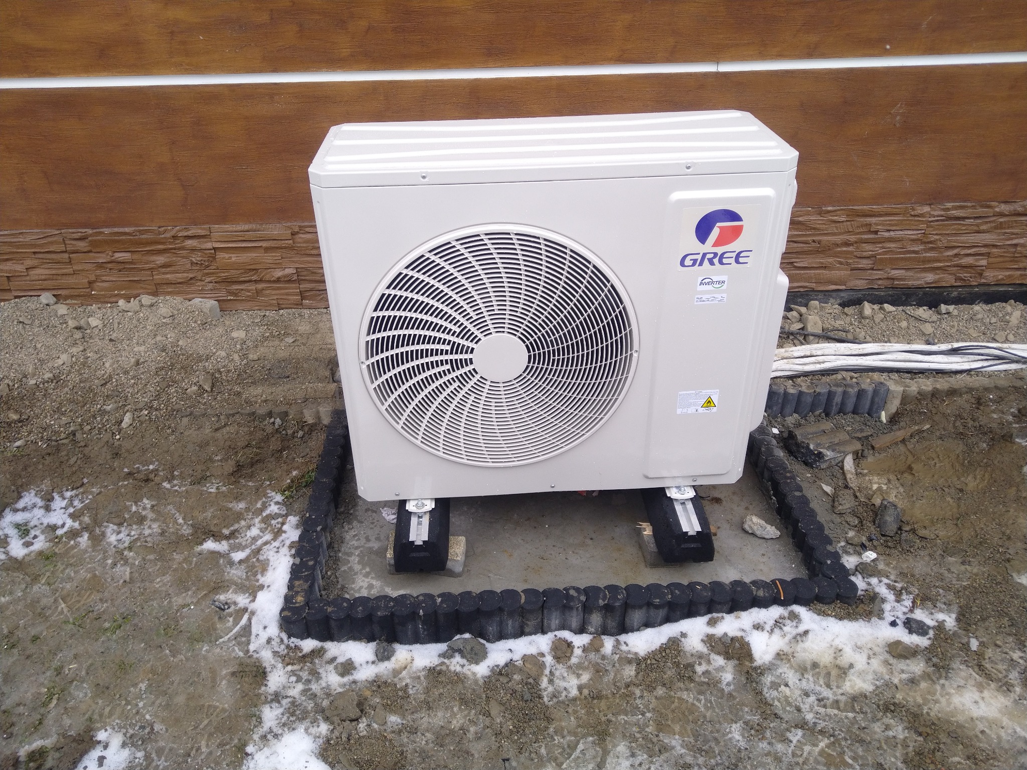 Zewnętrzna jednostka klimatyzatora marki Gree zamontowana na betonowych podstawach, otoczona czarnym obrzeżem, widoczne przewody i fragment elewacji budynku.