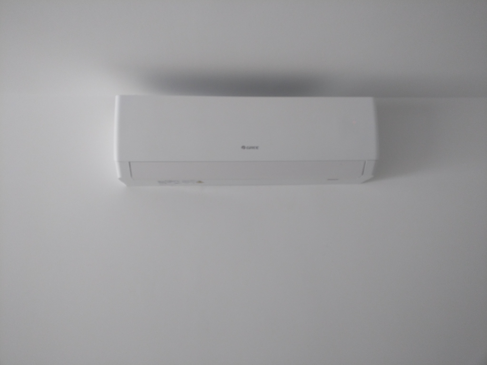 Biała jednostka wewnętrzna klimatyzacji Gree zamontowana na białej ścianie, widoczny cień na suficie.