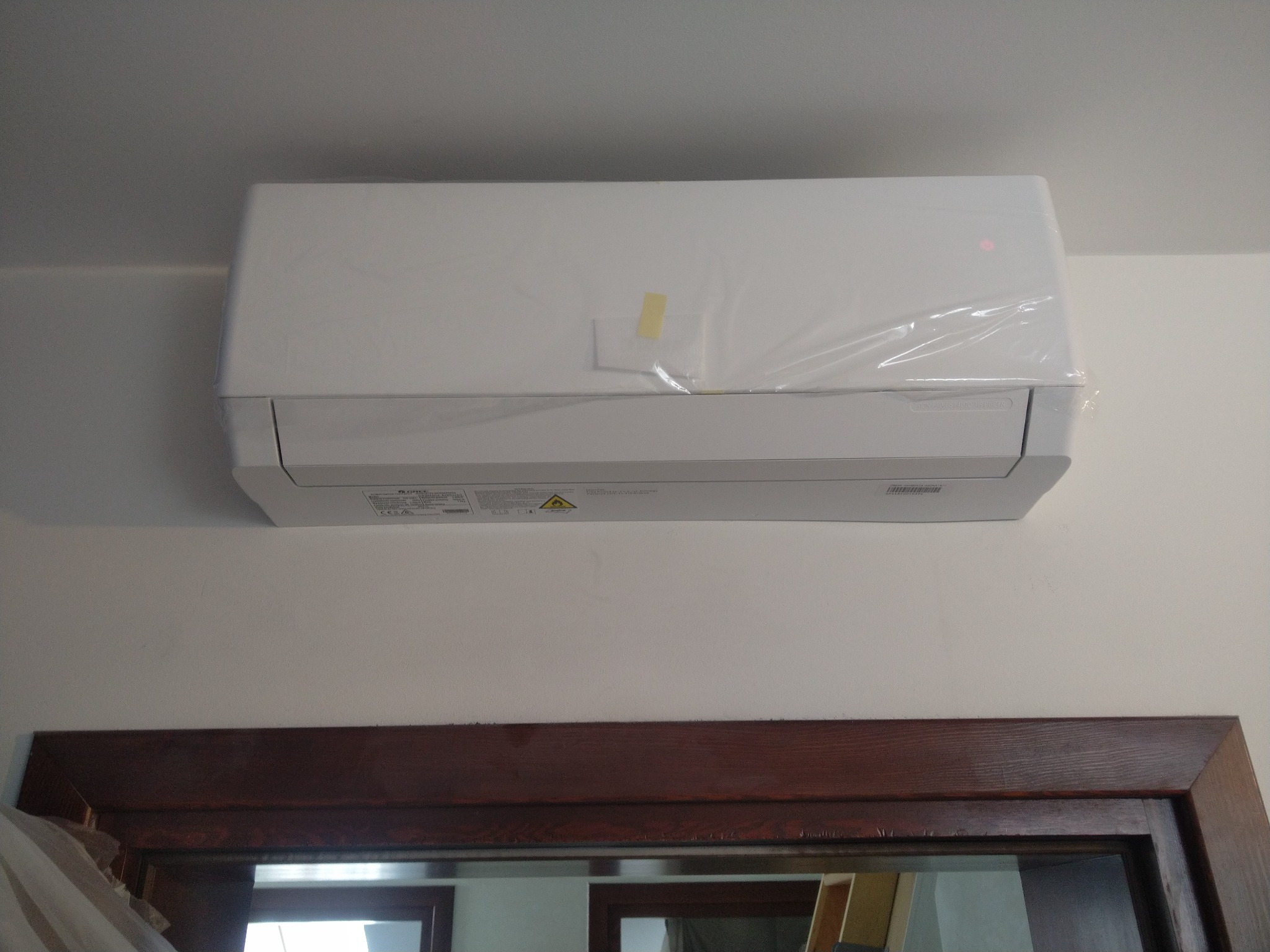 Nowy, biały klimatyzator ścienny, jeszcze w folii ochronnej, z widoczną taśmą zabezpieczającą, zamontowany nad ciemną, drewnianą framugą drzwi.