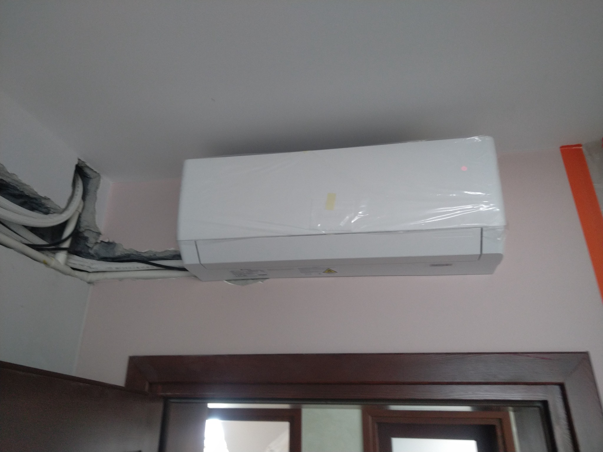 Nowo zamontowana, biała jednostka klimatyzacji wewnętrznej, jeszcze w folii ochronnej, widoczne przewody w ścianie i fragment futryny drzwi.
