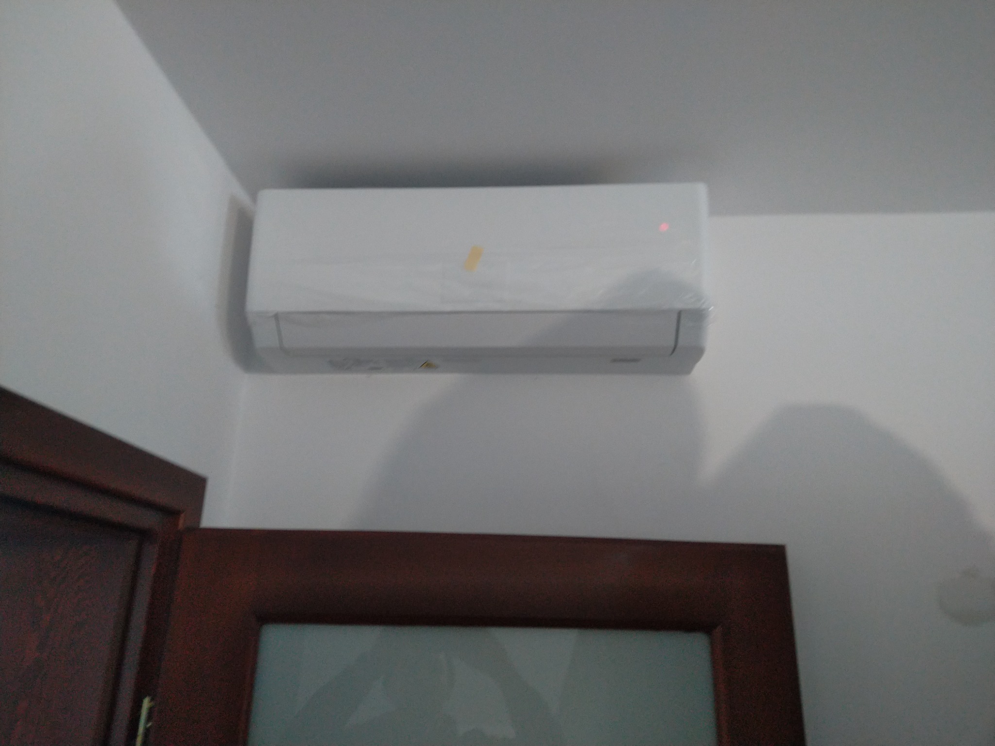 Biały klimatyzator ścienny zamontowany w rogu pokoju nad ciemnymi drzwiami z szybą, widoczna folia ochronna i czerwona dioda.