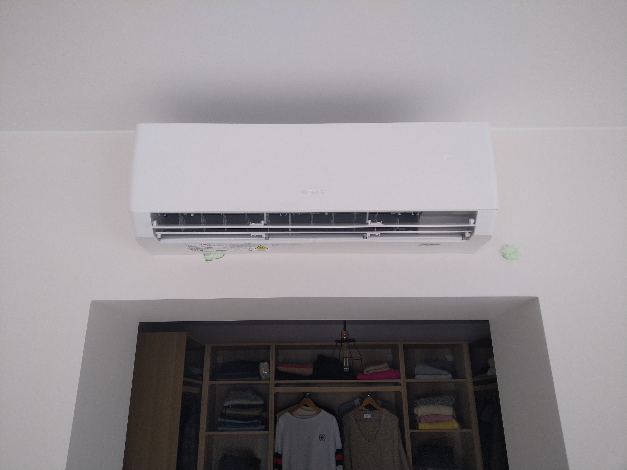 Biały klimatyzator marki Gree zamontowany nad wejściem do garderoby z widoczną zawartością półek i wieszakami z ubraniami.