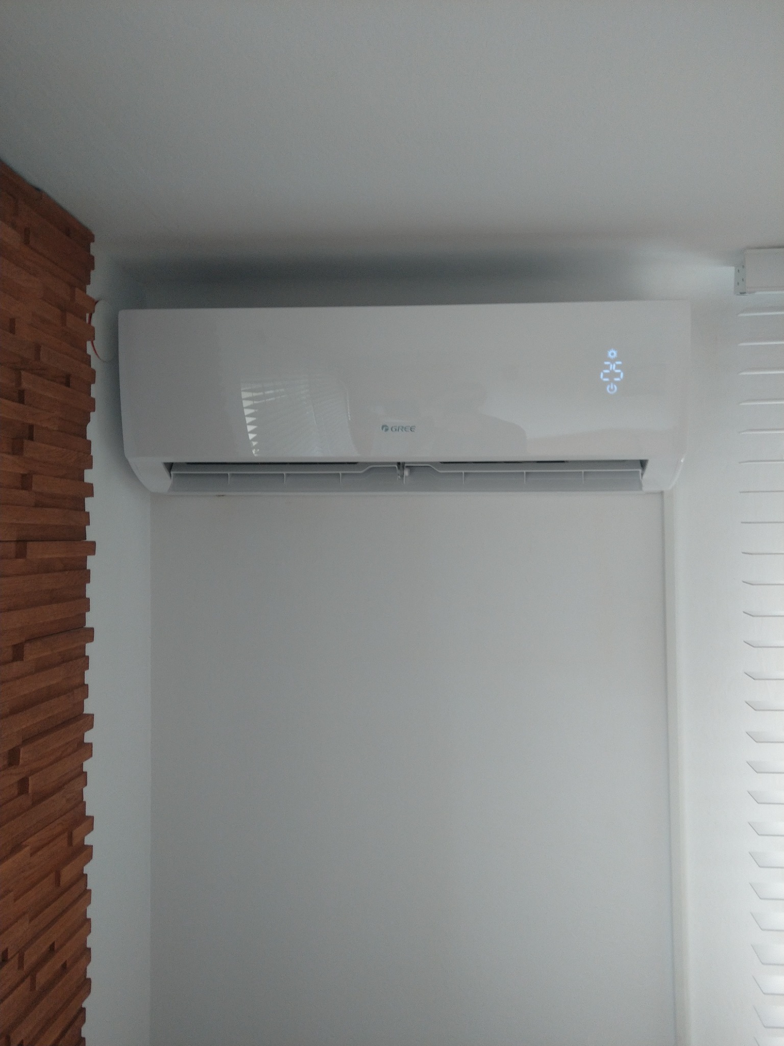 Biały klimatyzator marki Gree zamontowany na ścianie w pomieszczeniu, widoczny wyświetlacz temperatury 25 stopni Celsjusza, obok drewniana dekoracja ścienna i białe żaluzje.