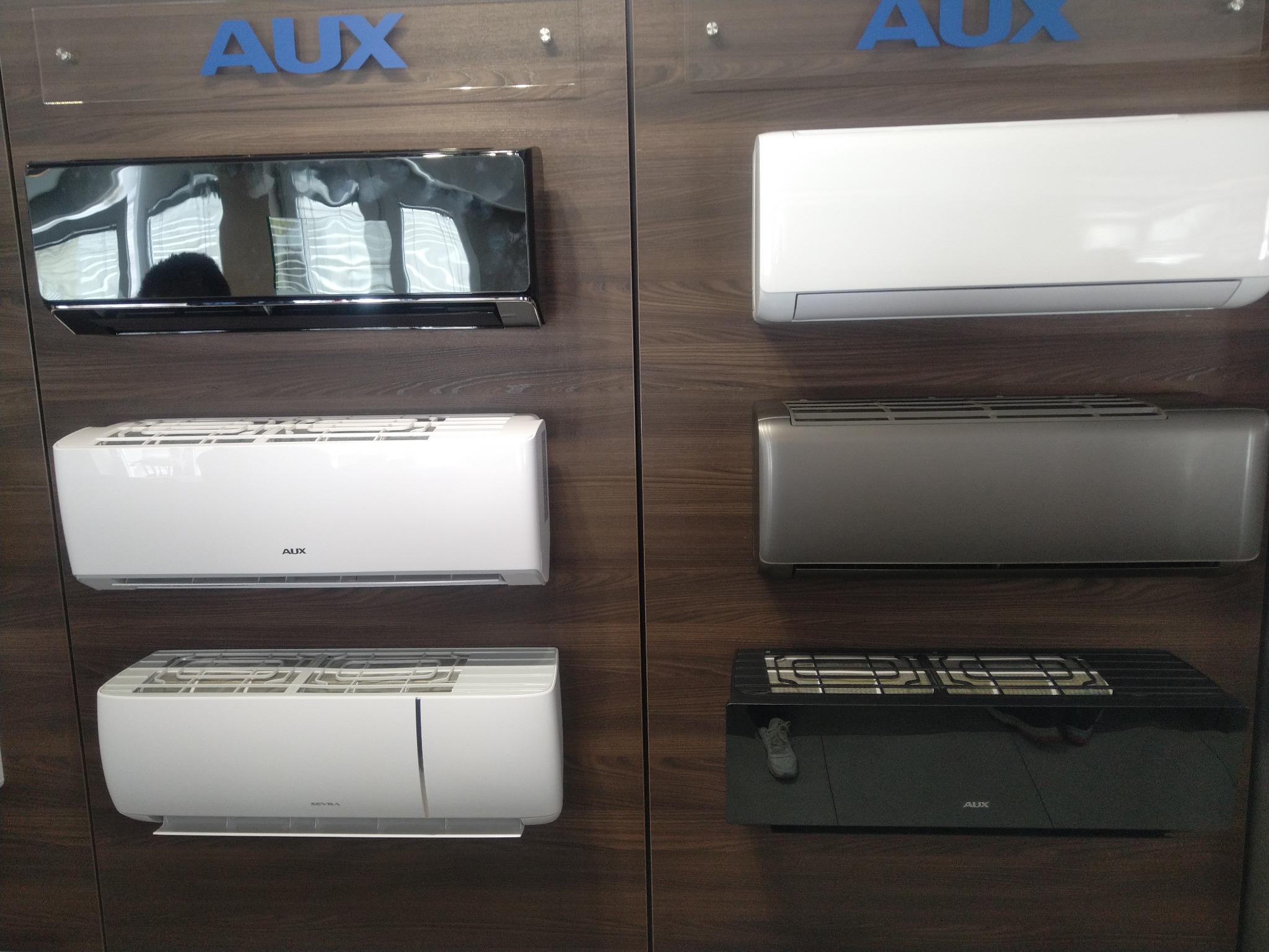 Ekspozycja różnych modeli klimatyzatorów marki AUX na drewnianej ścianie, widoczne modele w kolorze białym, srebrnym i czarnym.