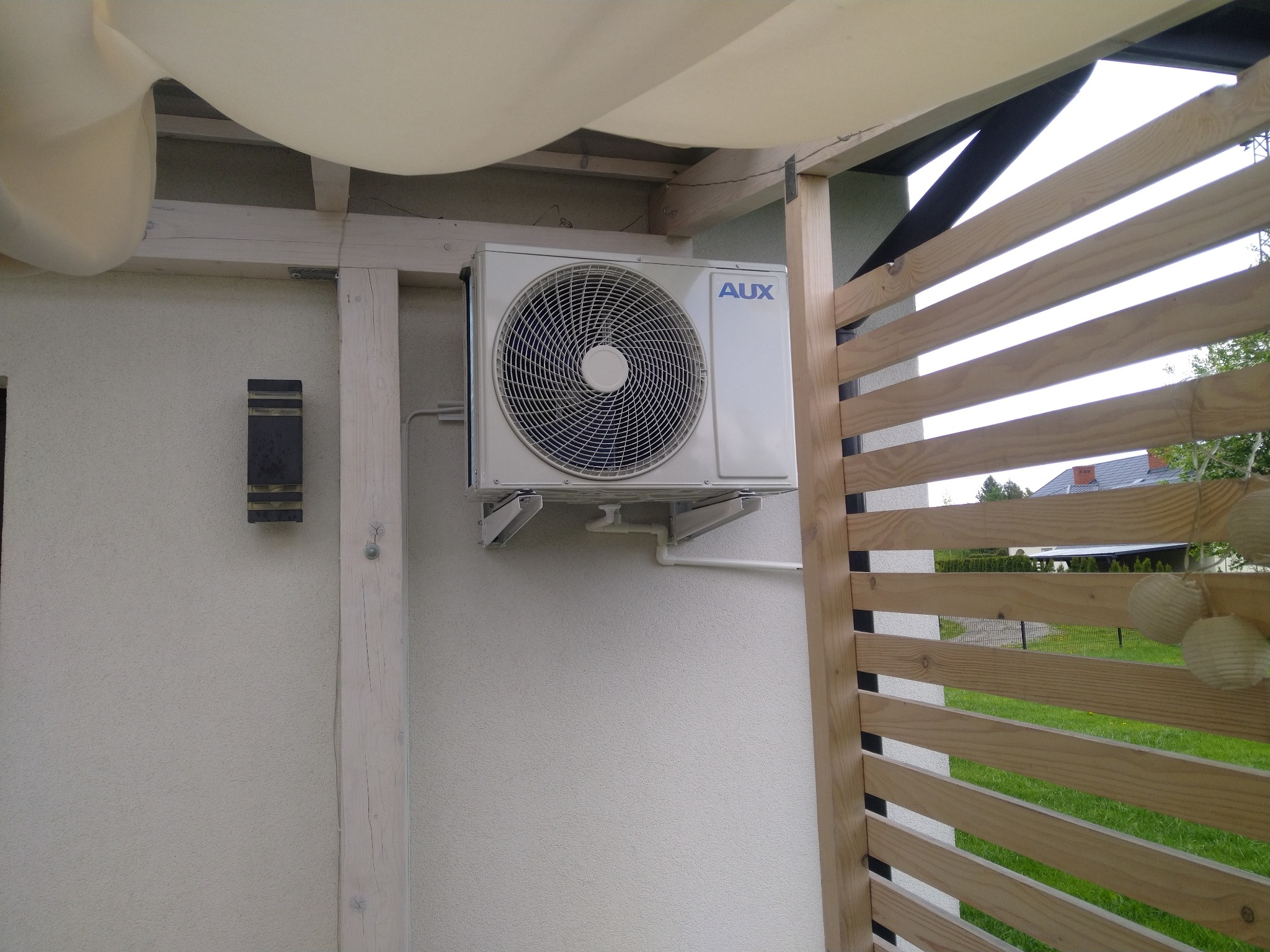 Zewnętrzna jednostka klimatyzacji AUX zamontowana na ścianie domu z drewnianą konstrukcją i markizą przeciwsłoneczną w tle.