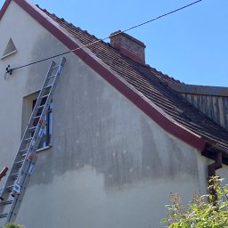 Szuru-Buru Robert Suchodolski - Ściana domu z widocznymi śladami po czyszczeniu, drabina oparta o ścianę, dach z czerwonymi obrzeżami, ceglany komin, błękitne niebo w tle.