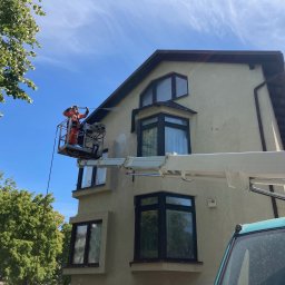 Szuru-Buru Robert Suchodolski - Pracownik w pomarańczowym kombinezonie na podnośniku koszowym czyści elewację dwupiętrowego domu strumieniem wody pod ciśnieniem, widoczny fragment nieumytego i umytego muru.